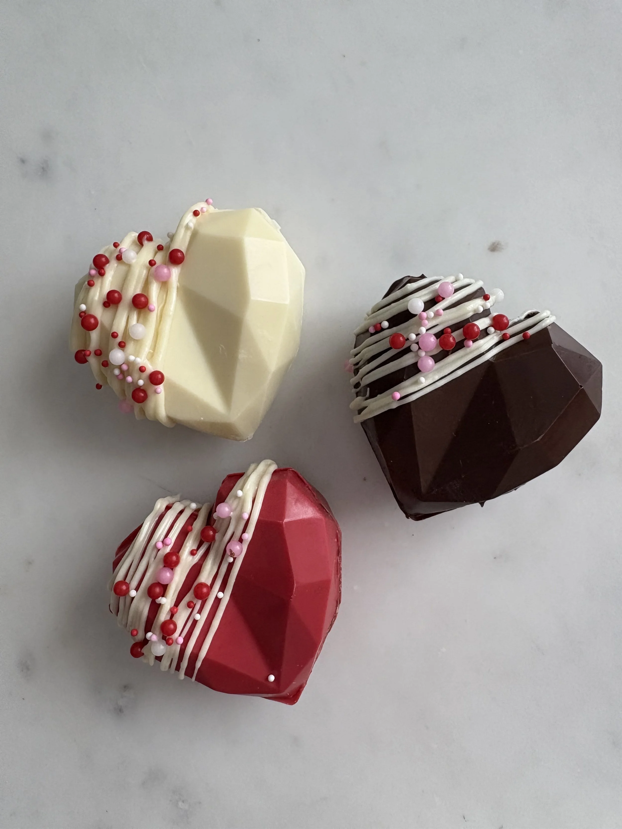 Three heart-shaped chocolates with white, red, and pink sprinkles and drizzle decorated on top, placed on a white surface.