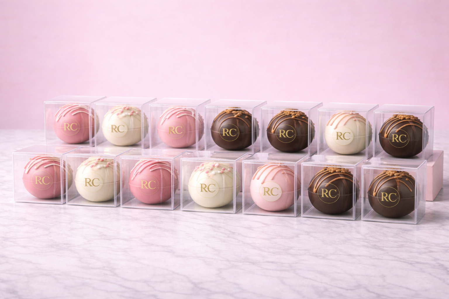 Assorted decorated chocolates in clear boxes with gold 'RC' logos, arranged on a marble surface against a pink background.