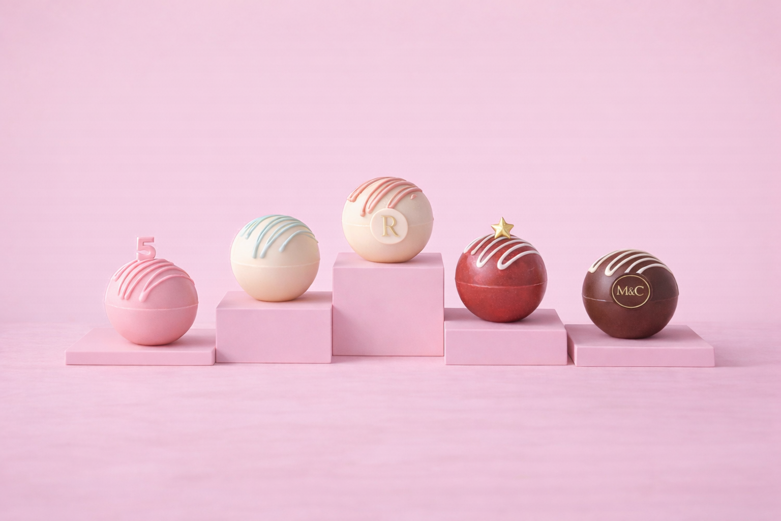 Five decorated spherical chocolates placed on pink display platforms against a pink background, each with unique designs and markings.