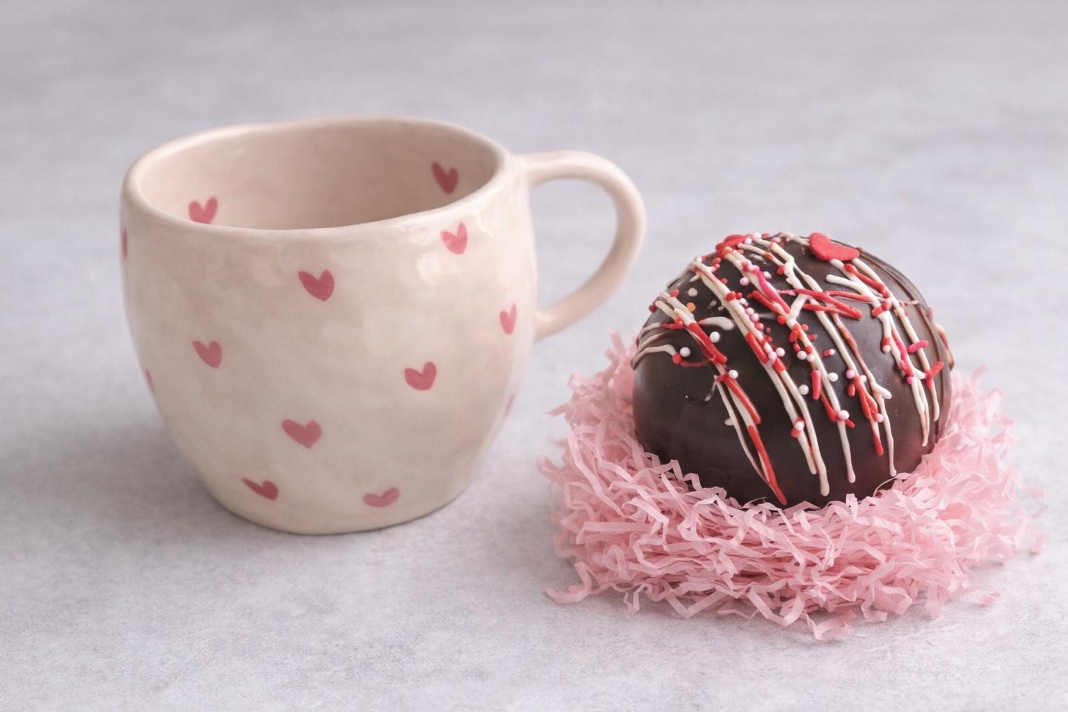 A ceramic mug with pink hearts and a decorated chocolate truffle sitting on pink shredded paper.