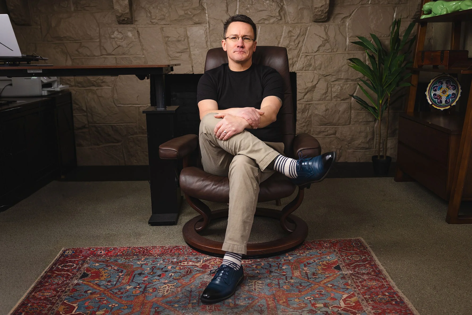 A man sitting in an office chair with crossed legs, wearing a black t-shirt, beige pants, striped socks, and black shoes, in a room with a stone wall, a wooden bookshelf, a green plant, and a red patterned rug.