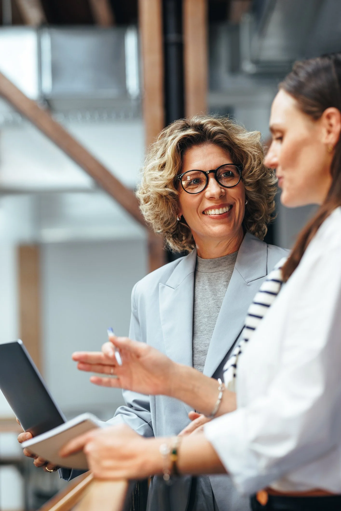 Two women are in discussion in an office setting, with one woman smiling and holding a tablet, and the other woman gesturing with her hand, holding a notepad.