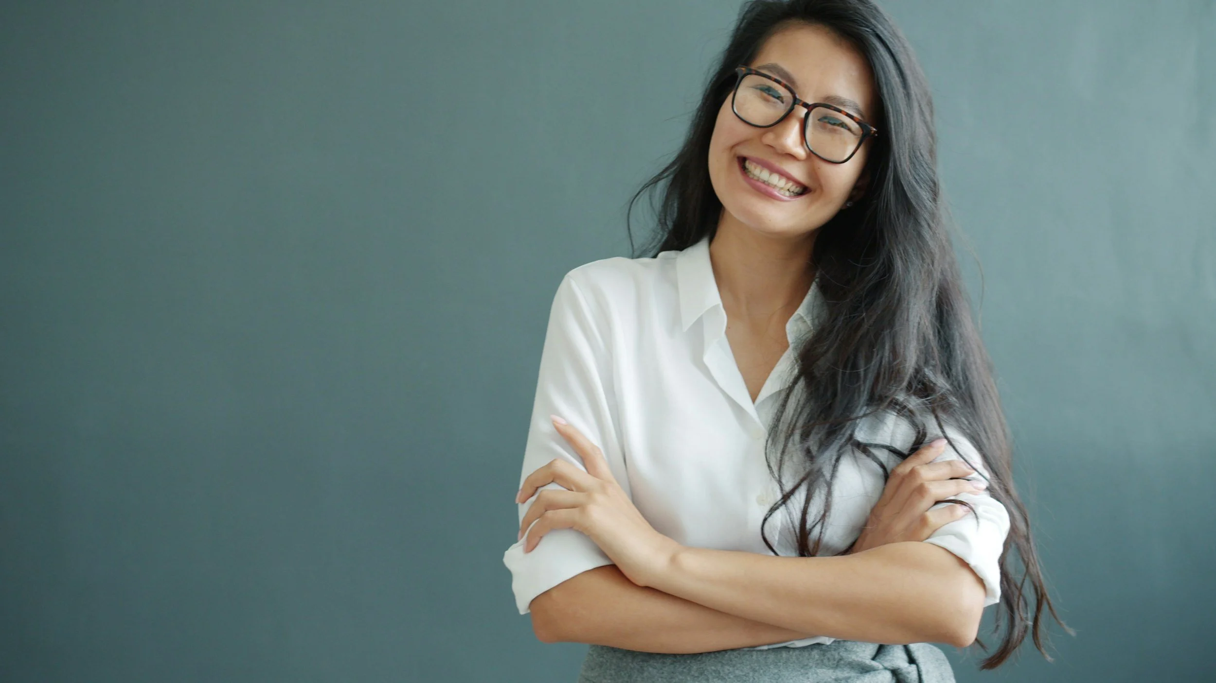 Smiling woman with long dark hair, glasses, white shirt, arms crossed, standing against a plain wall.