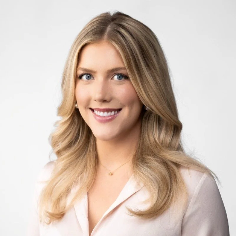 A woman with blonde hair and blue eyes smiling, wearing a light-colored blouse against a plain background.