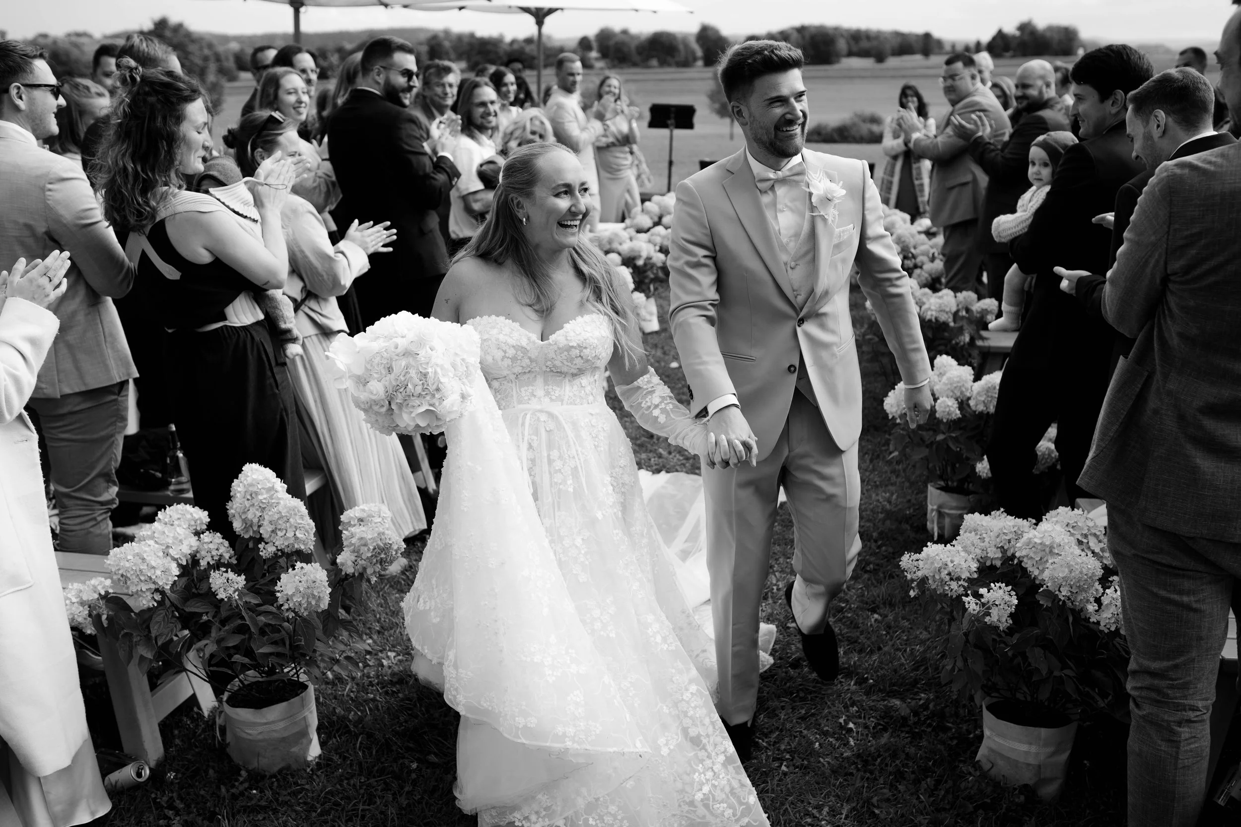 Braut und Bräutigam halten Händchen während ihrer Hochzeit, umgeben von lachenden Gästen im Freien.