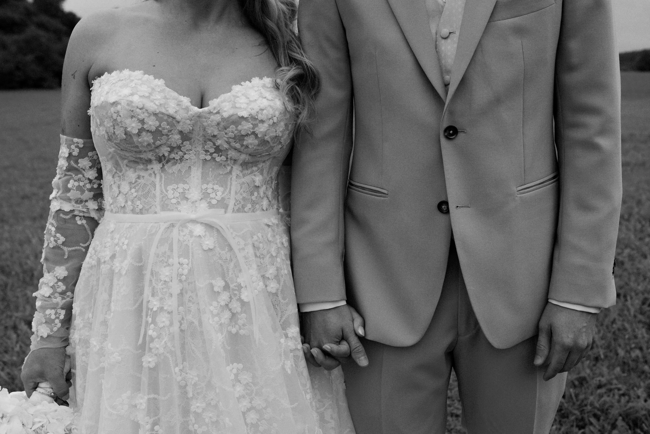 Ein Brautpaar hält sich an den Händen, die Braut trägt ein Hochzeitskleid mit Blumenmuster, der Bräutigam einen Anzug, sie stehen im Freien auf einer Wiese.