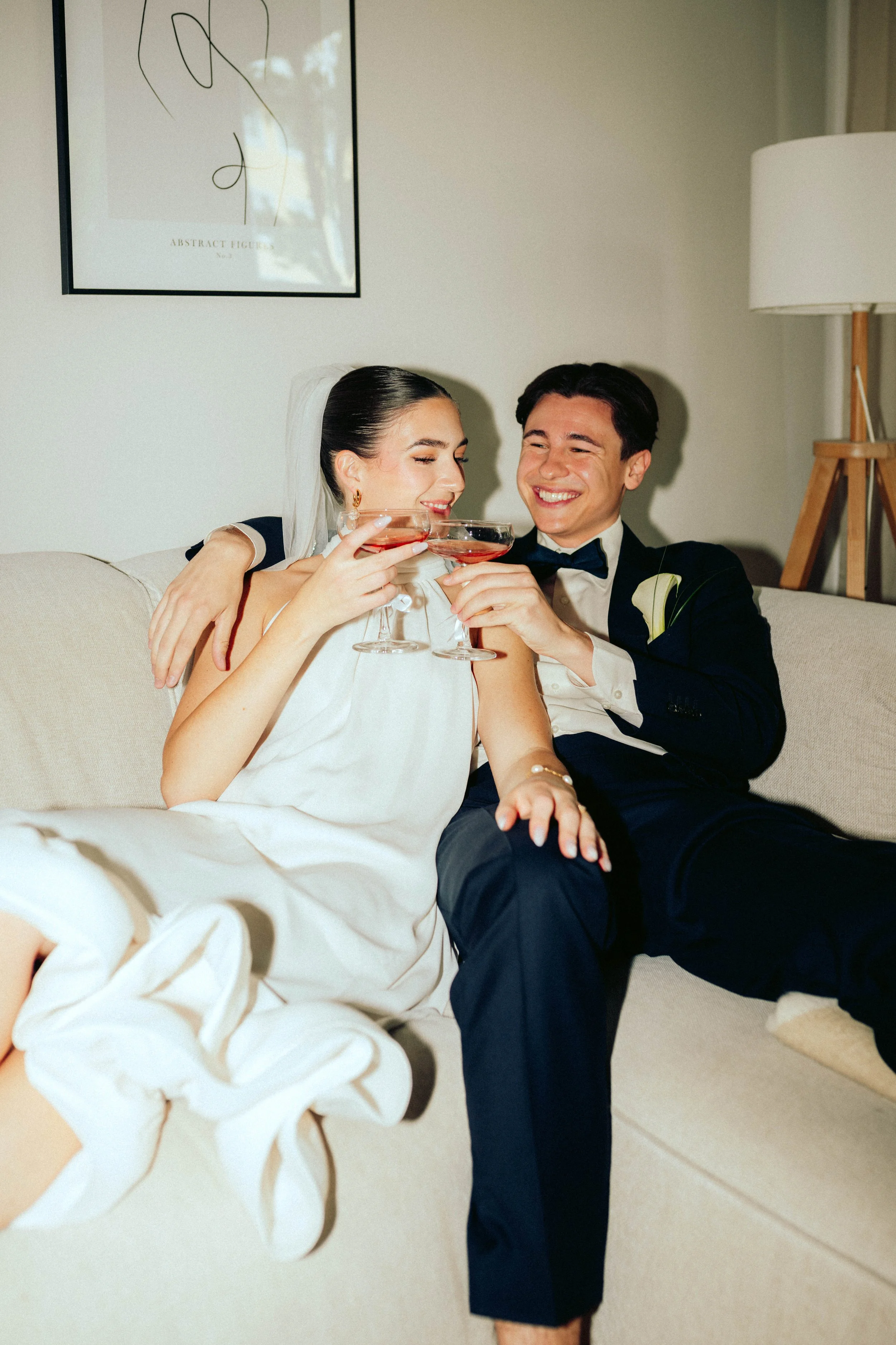 Ein lachendes Brautpaar sitzt auf einem Sofa, trinkt auf Hochzeitsfeier Wein, und trägt formelle Kleidung, der Mann in einem schwarzen Anzug mit Fliege, die Frau in einem weißen Brautkleid mit Schleier.