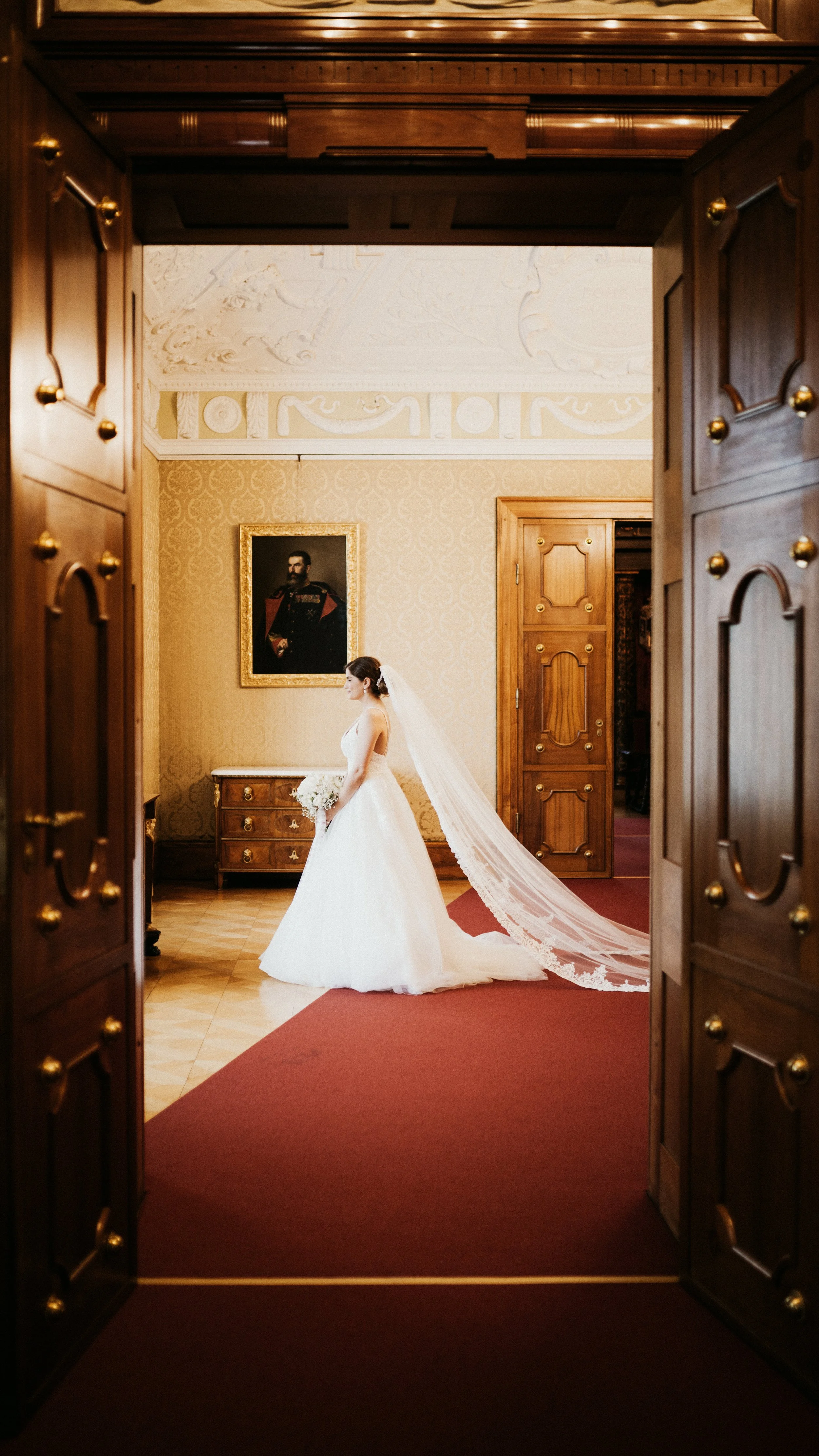 Braut in einem weißem Hochzeitskleid mit Schleier steht in einem eleganten Raum, Blick nach links, im Hintergrund eine historische Gemäldeporträt an der Wand, Holzvertäfelung und rote Teppichboden.