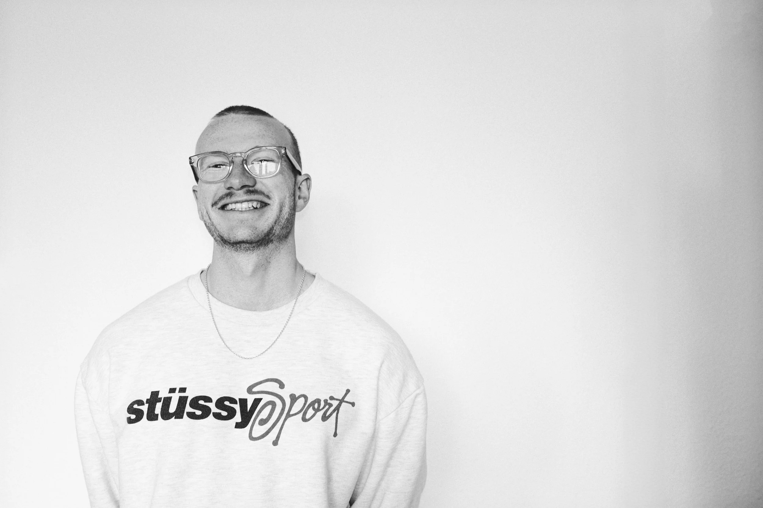 Junger Mann mit Brille, lächelnd, trägt ein Stüssy Sport Sweatshirt, steht vor einer hellen Wand.