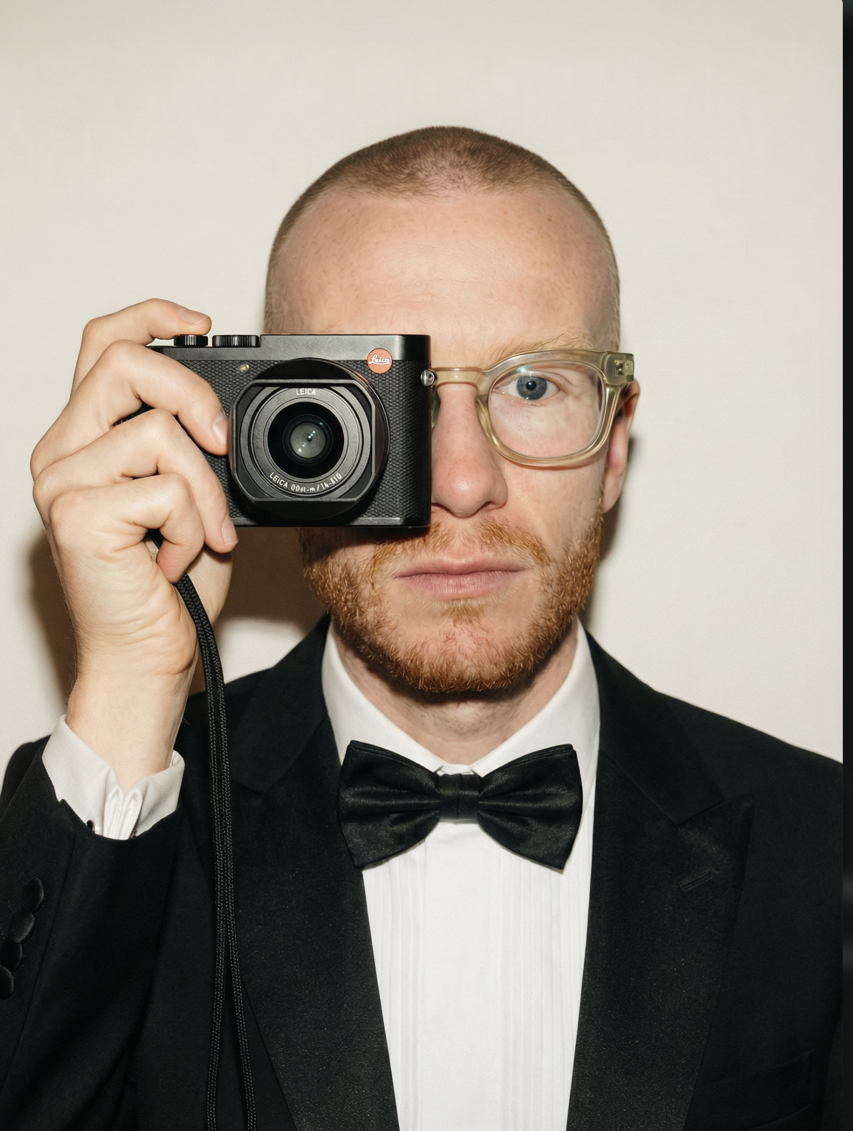 Mann in formeller Kleidung hält Kamera vor sein linkes Auge, trägt Brille, dunkler Anzug, weißes Hemd, schwarze Fliege, kurz geschnittenes Haar, rotes Bart, neutraler Gesichtsausdruck gegen hellen Hintergrund.