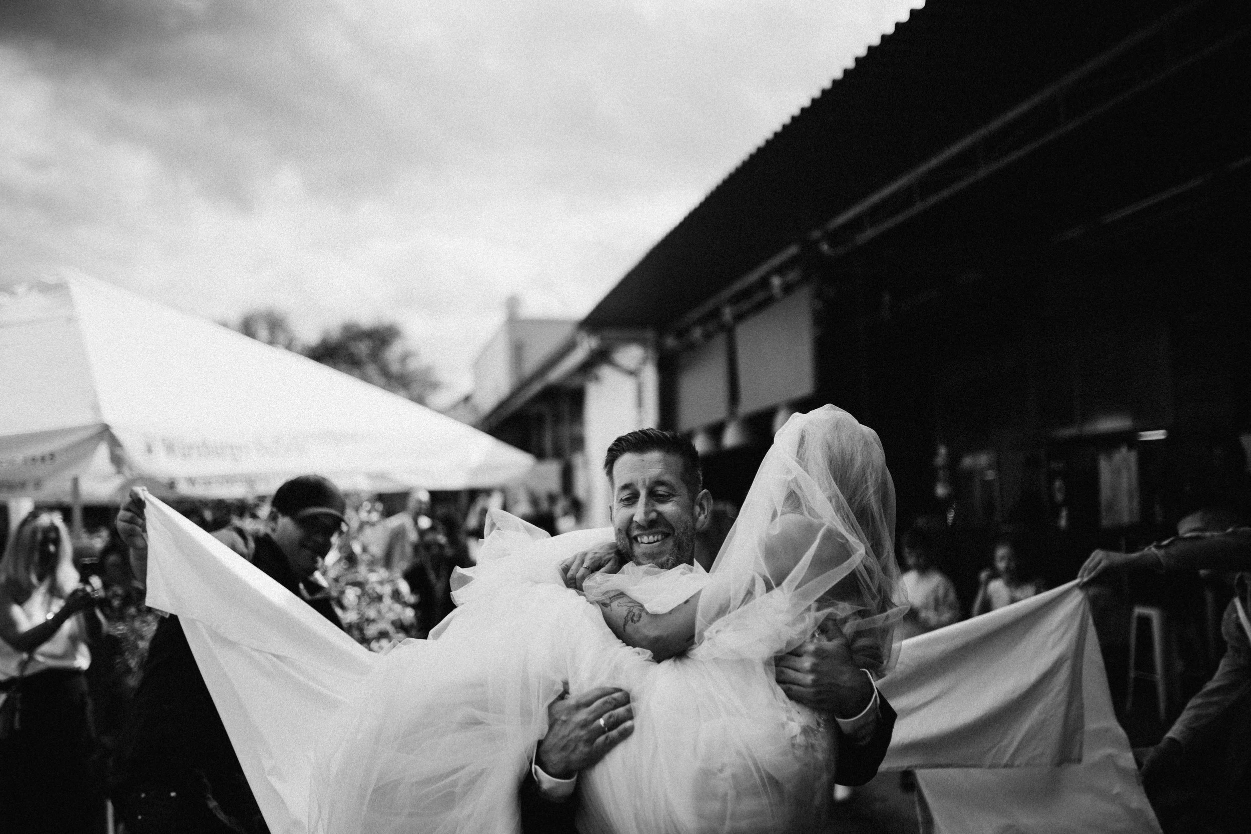 Ein Mann hält eine lächelnde Frau in einem weißen Kleid bei einer Feier im Freien, umarmt sie und trägt sie hoch. Es sind weitere Menschen im Hintergrund.