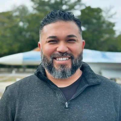 Smiling man with dark hair and beard, wearing a black zip-up jacket outdoors with trees and a boat in the background.
