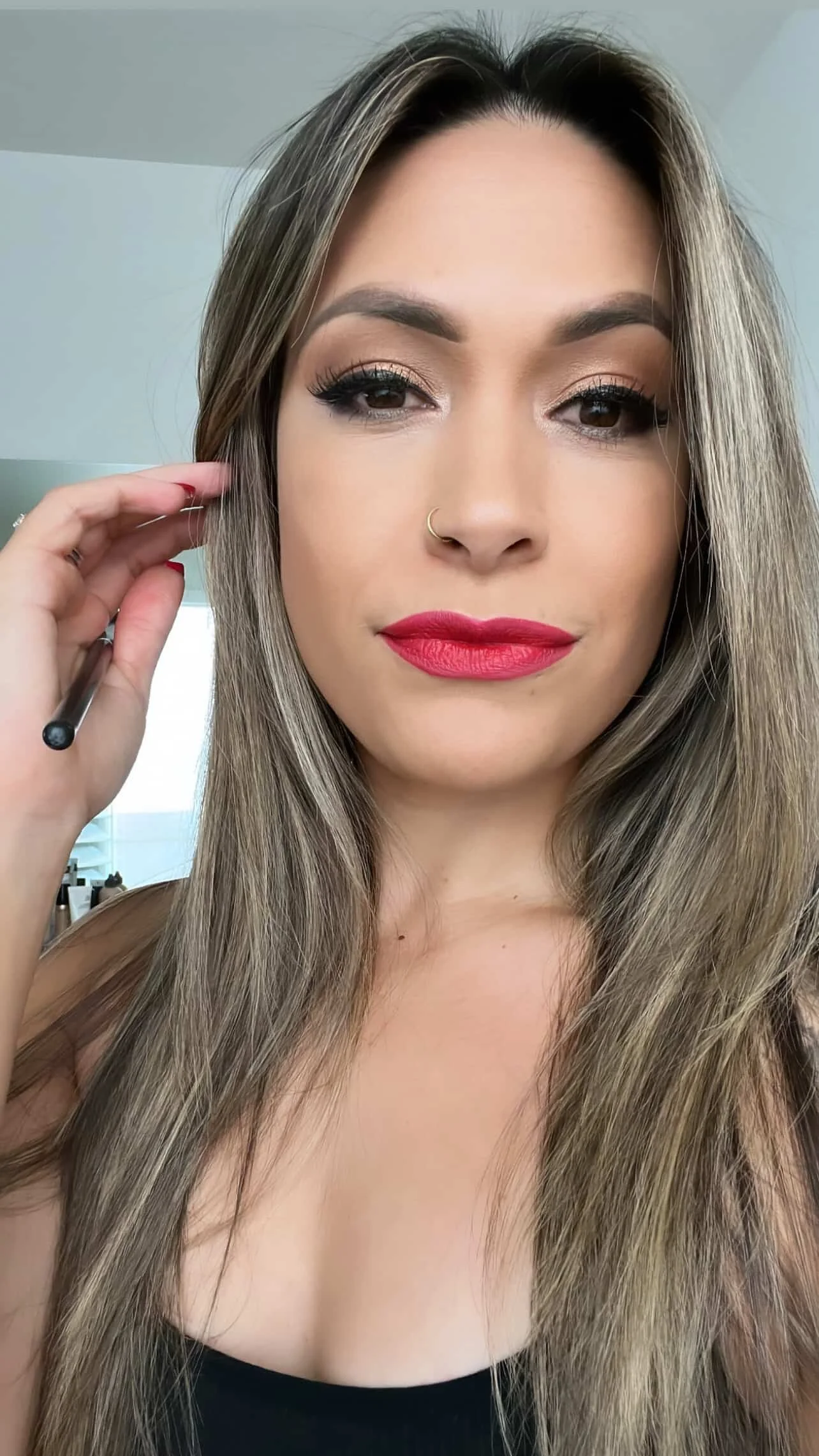 A woman with long brown hair, wearing makeup, a gold nose ring, and red lipstick, holding a makeup brush near her face.