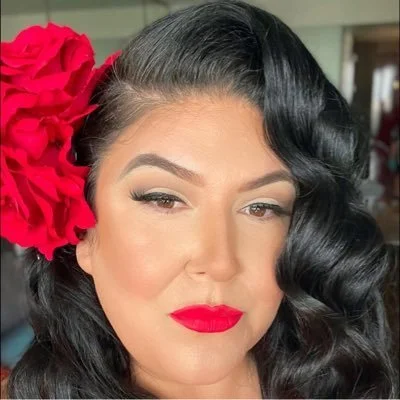 Woman with dark wavy hair, red lipstick, red hair accessory, and makeup, looking at the camera.