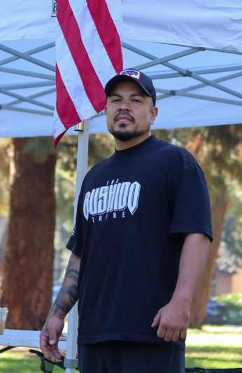 Man wearing a black Rushdo Tribe shirt and baseball cap standing outdoors under a tent with an American flag in the background.