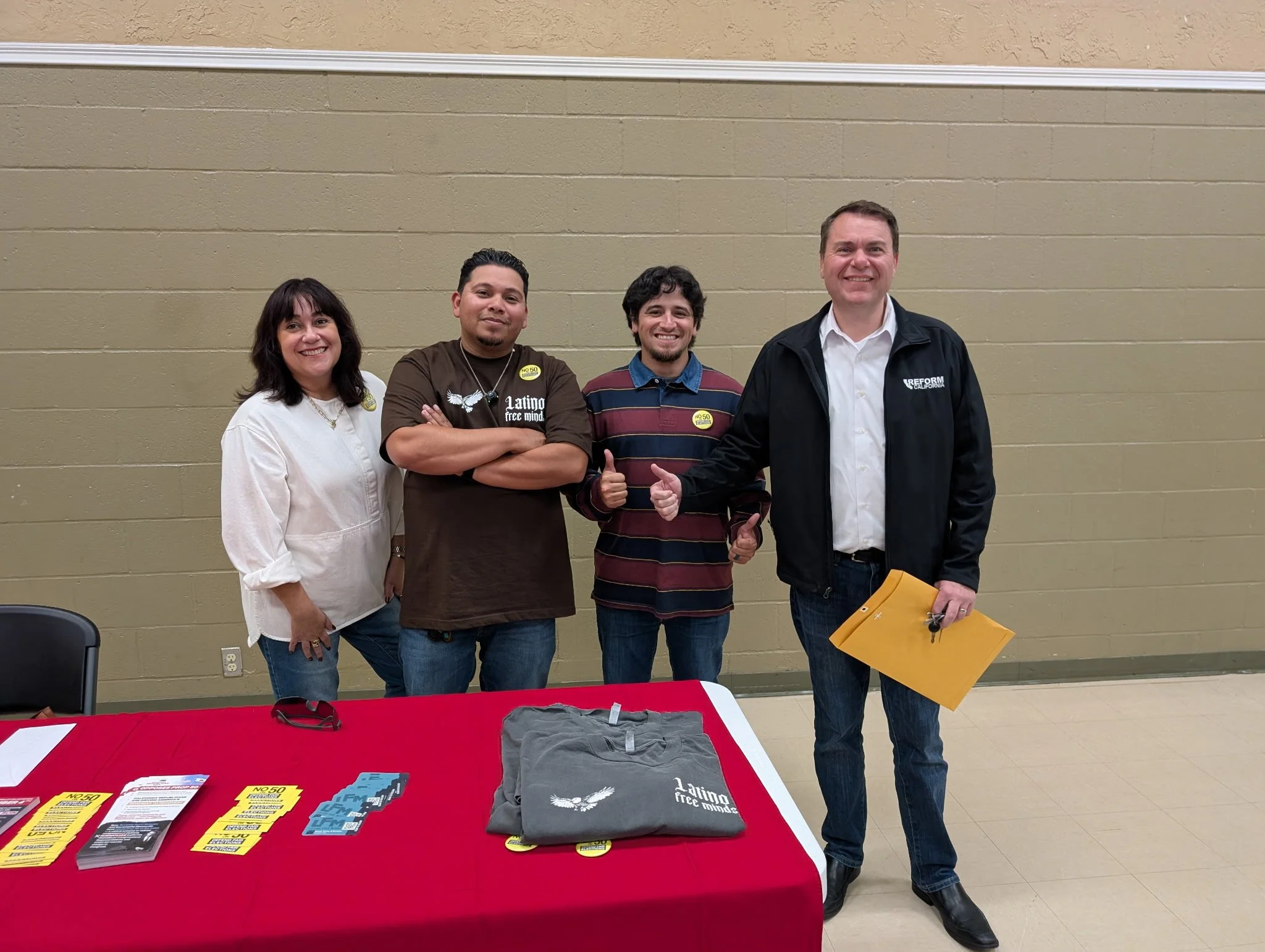Four people standing behind a table covered with promotional materials, smiling and giving thumbs up, against a beige wall.