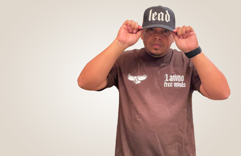 Man wearing a black cap with the word 'lead', adjusting it with both hands, standing against a plain background.