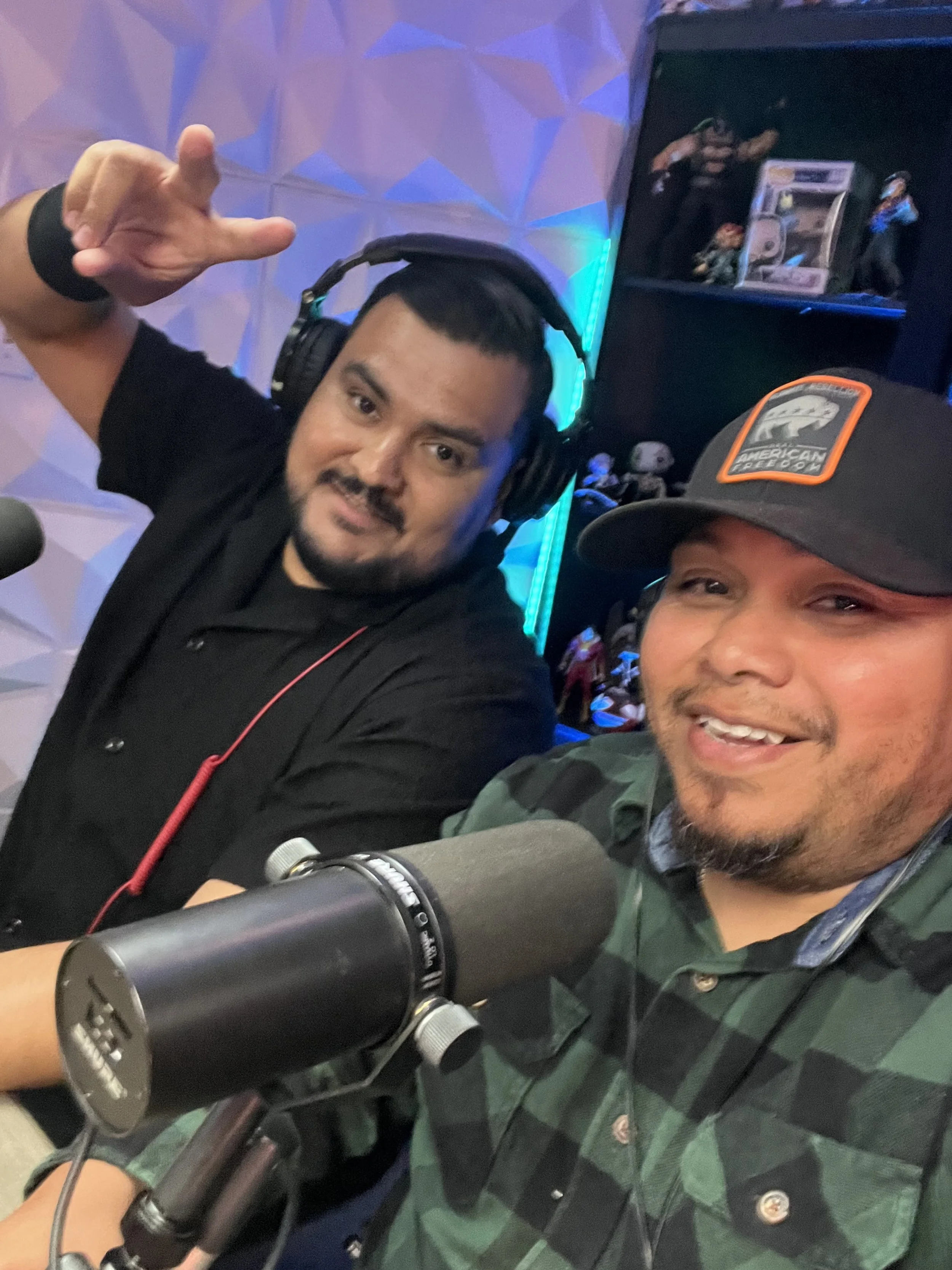 Two men taking a selfie in a recording studio, one wearing headphones and the other with a microphone in front of him. The man with the microphone is smiling, and the other is making a hand gesture.