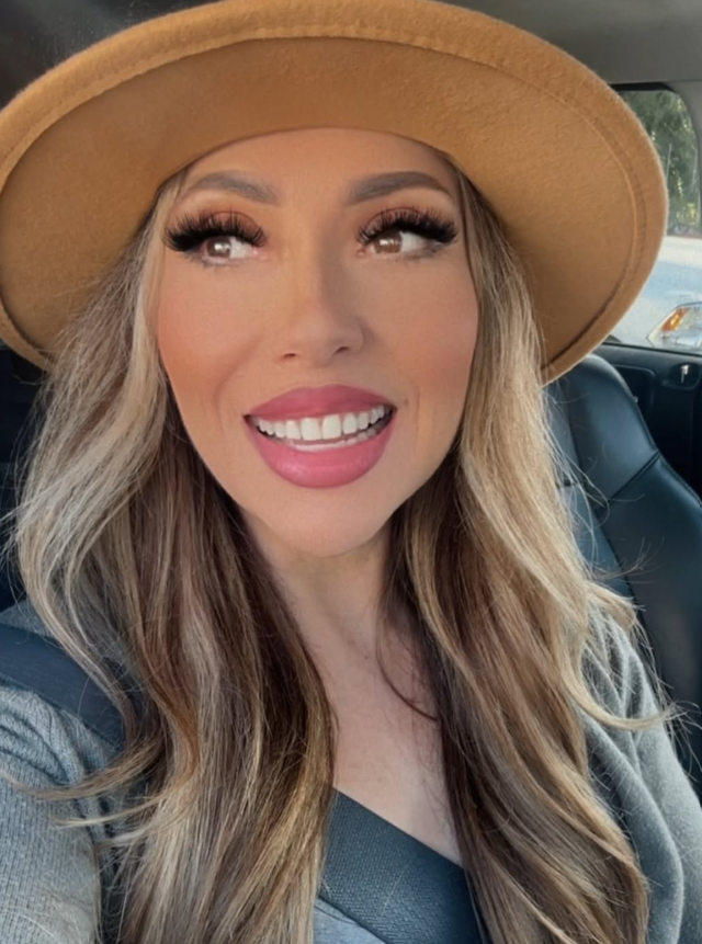 Woman with blonde wavy hair, makeup, and lipstick, smiling, wearing a tan hat and gray top inside a vehicle.