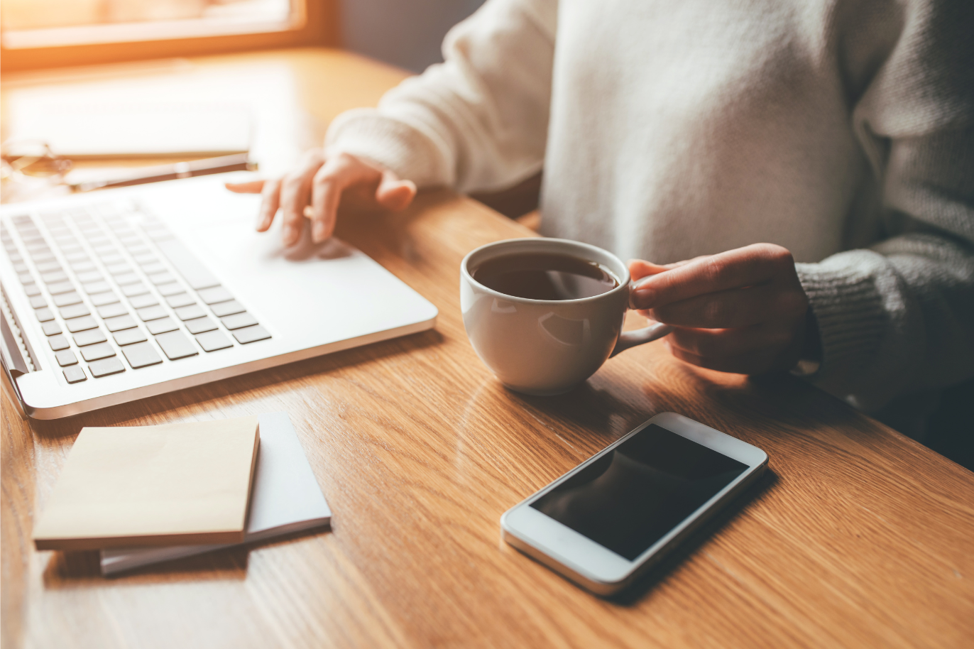 Personne travaillant à un bureau en utilisant un ordinateur portable, tenant une tasse de café, avec un smartphone et un bloc-notes à portée de main, dans un environnement lumineux.