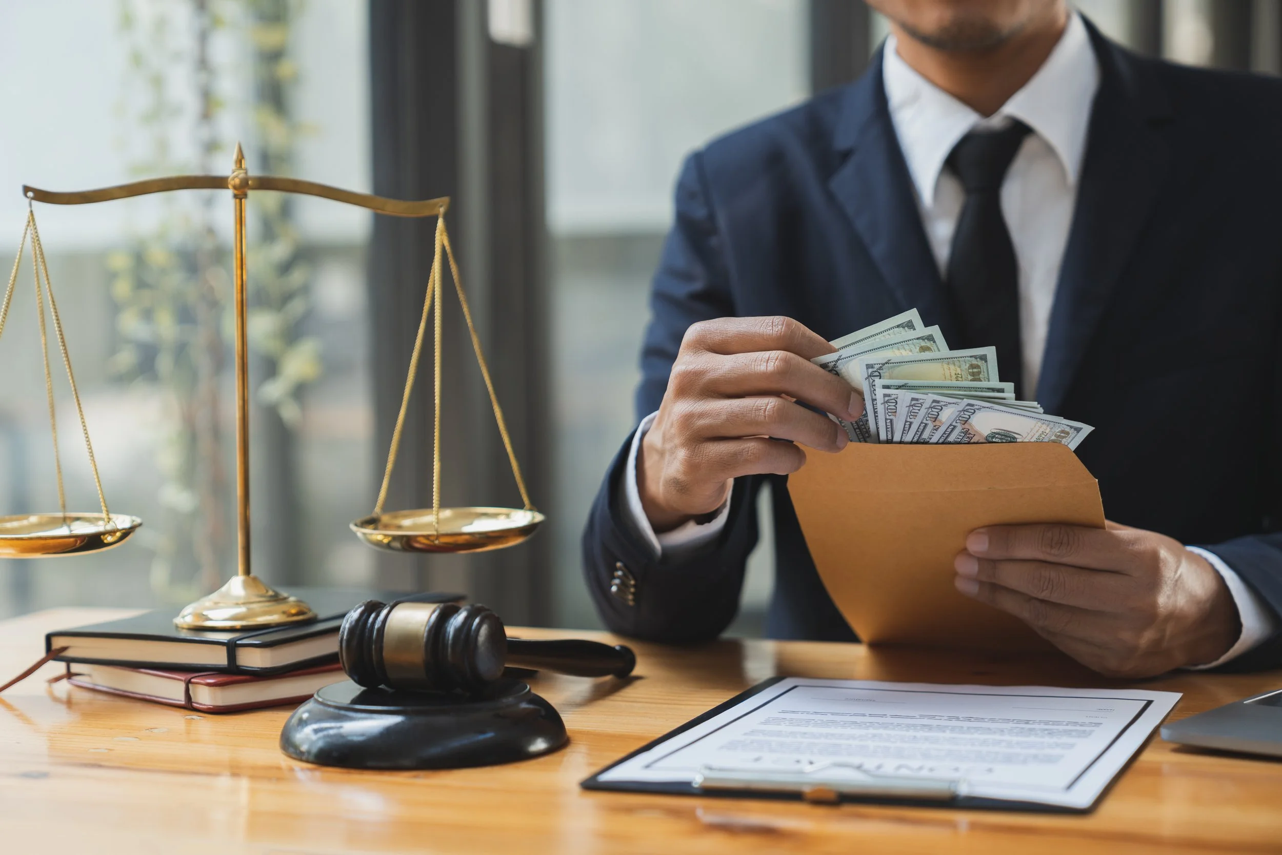 A man in a suit seated at a wooden desk, holding a folder filled with hundred-dollar bills, with a scale, gavel, books, and a legal document on the desk.