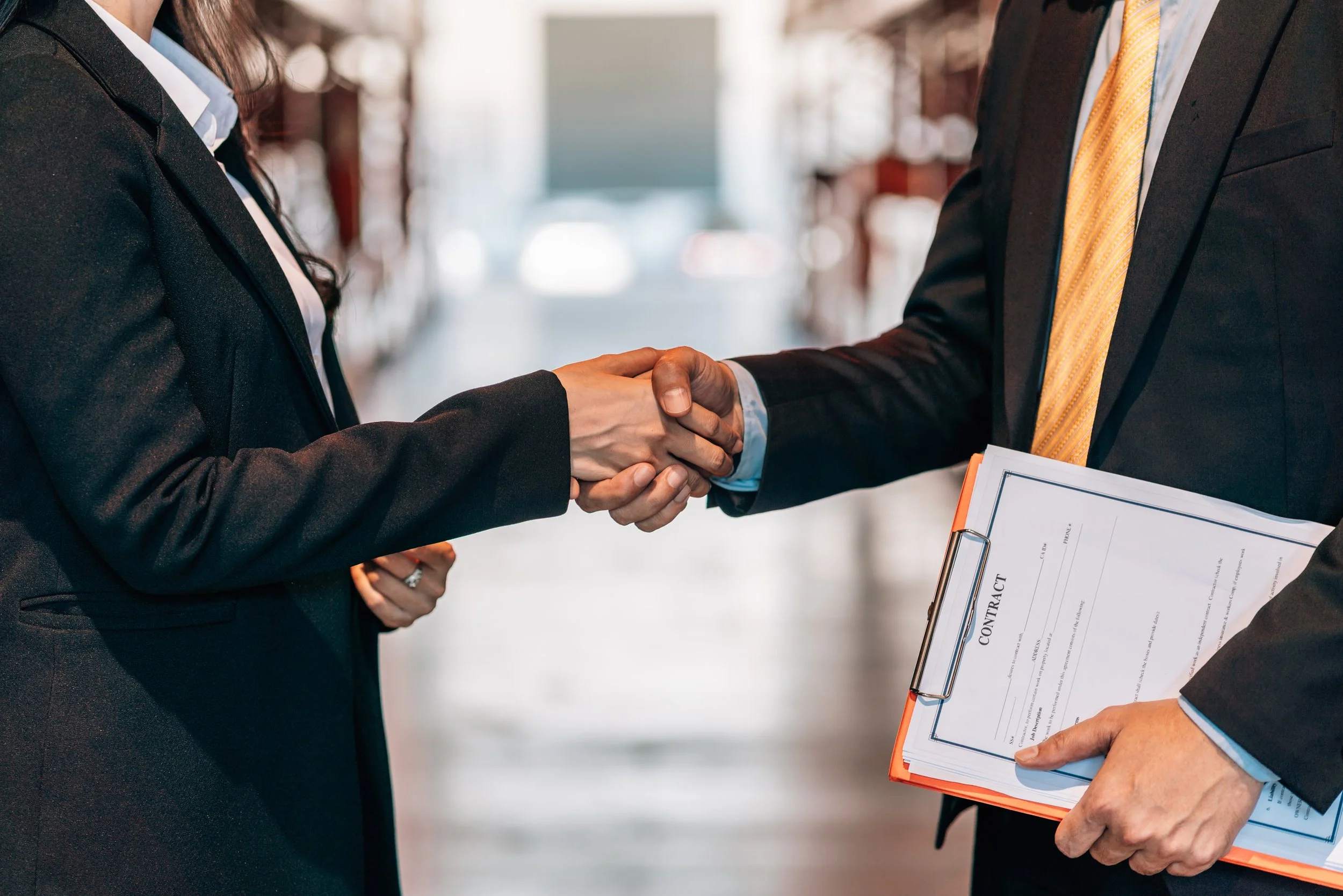 Two people in business suits shaking hands, one holding a clipboard with a contract, in a warehouse or storage facility.