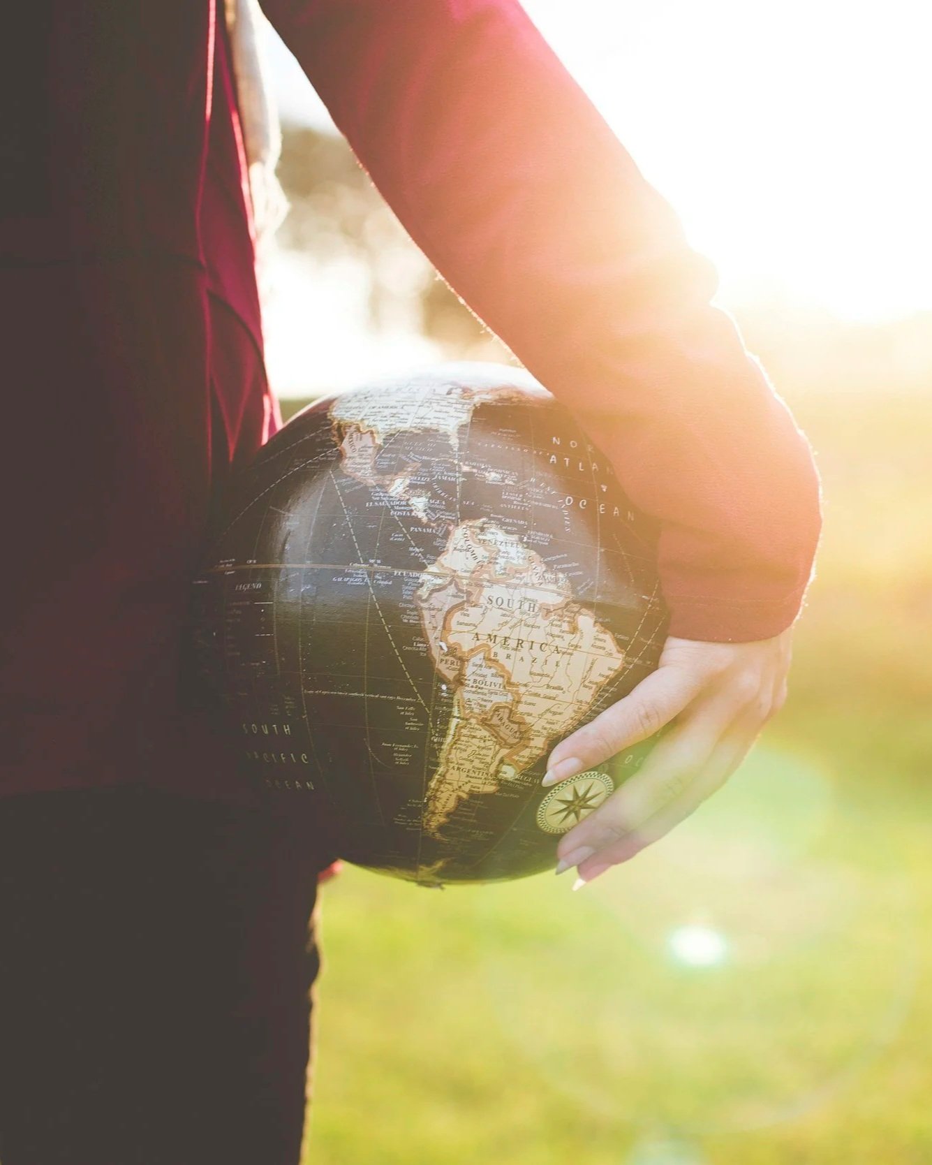 A person in a red jacket holding a globe showing South America outdoors during the daytime with sunlight shining from behind.