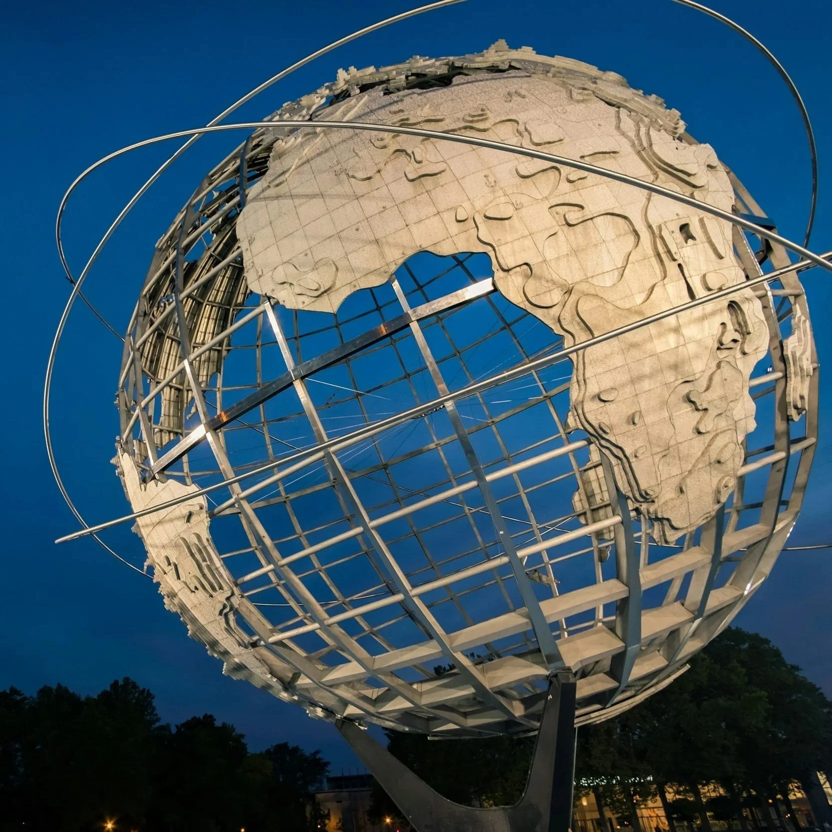 A large globe sculpture representing the Earth, made of metal and wood, showing continents carved in wood, with metal rings orbiting around it, illuminated against a blue evening sky with trees and lights below.