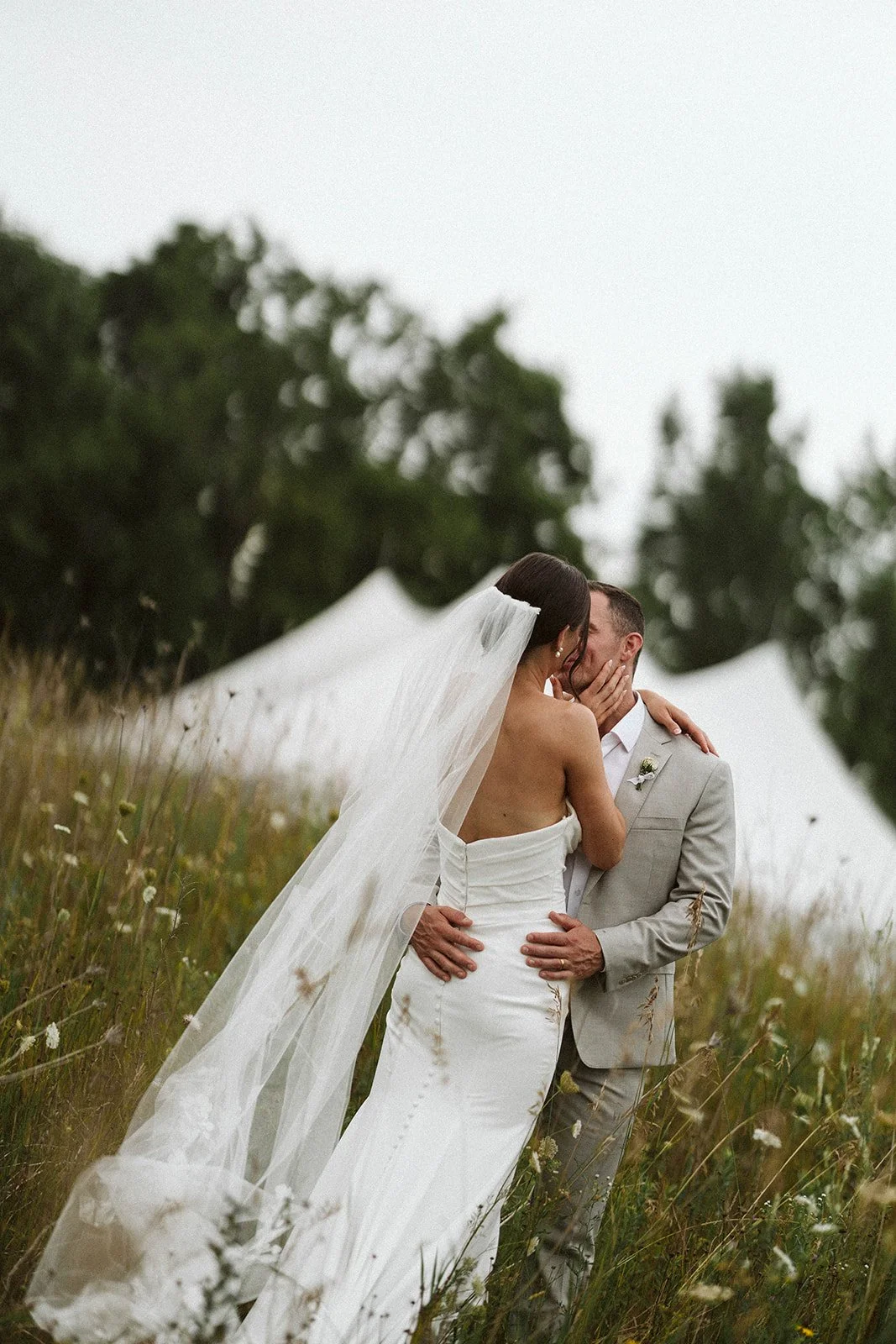 A bride and groom sharing a kiss in a grassy field during their outdoor wedding, with white tents in the background.