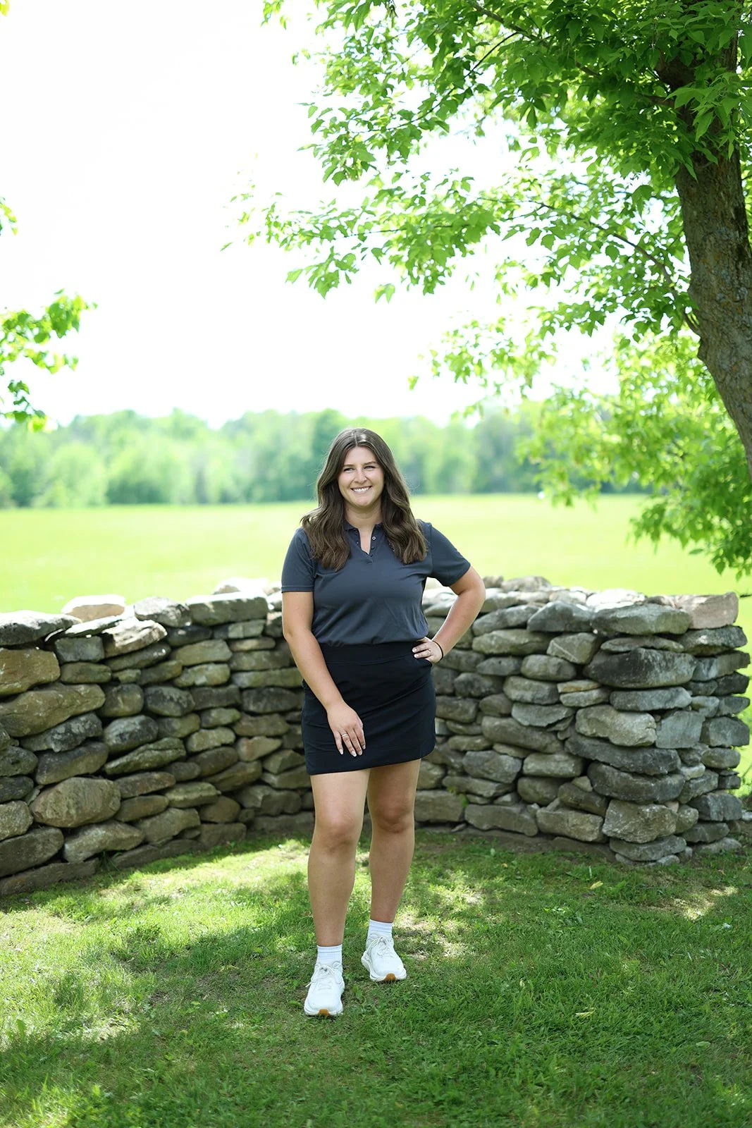 A woman standing outdoors near a stone wall, wearing a navy polo shirt, black skirt, white sneakers, and white socks, with green trees and open field in the background.