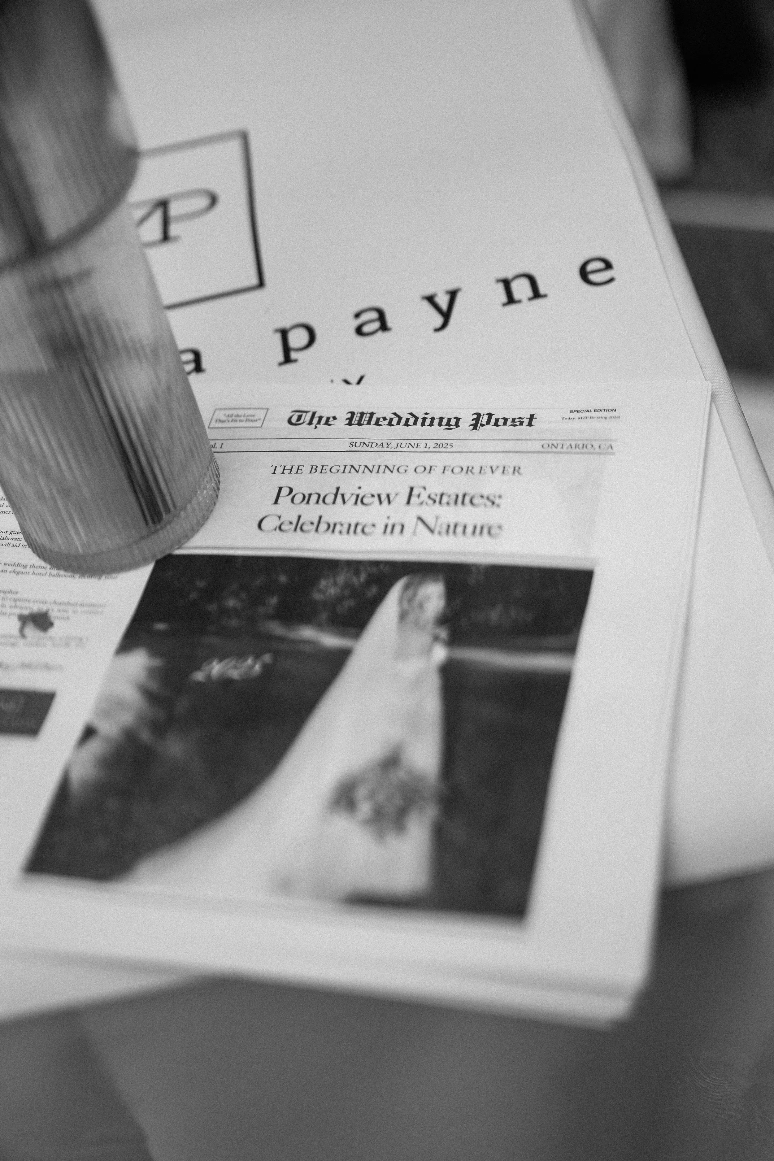 A newspaper titled 'The Wedding Post' with the headline 'Pondview Estates: Celebrate in Nature,' a photograph of a natural landscape, and some overlapping papers and a cylindrical container on top of the papers.