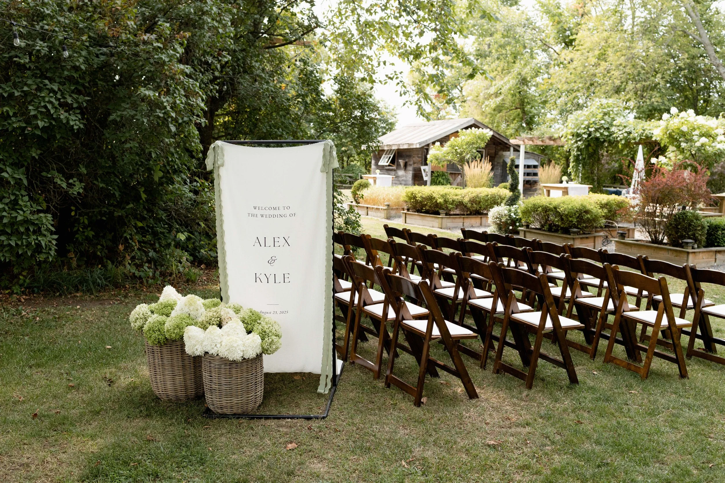 Outdoor wedding ceremony setup with wooden chairs arranged on grass, a welcome sign for the wedding of Alex and Kyle with white floral arrangements, greenery, and a rustic wooden building in the background.