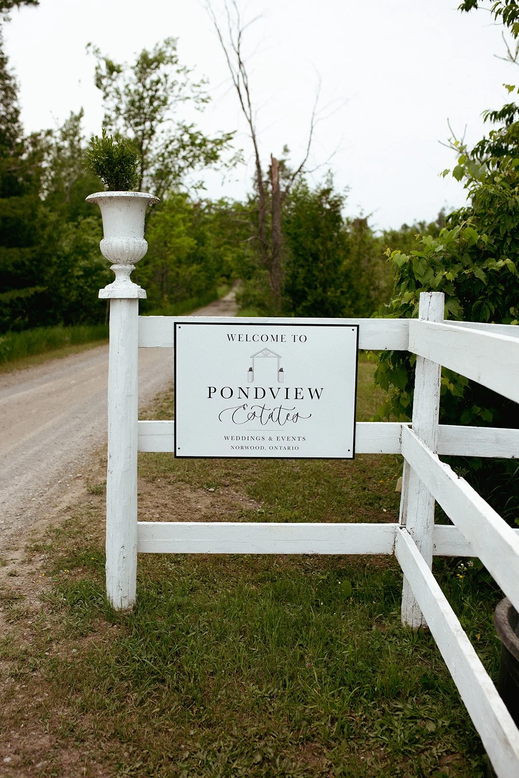 White signboard on a white fence welcoming visitors to Pondview Cottage for weddings and events in Norwood, Ontario, with a gravel path and green trees in the background.