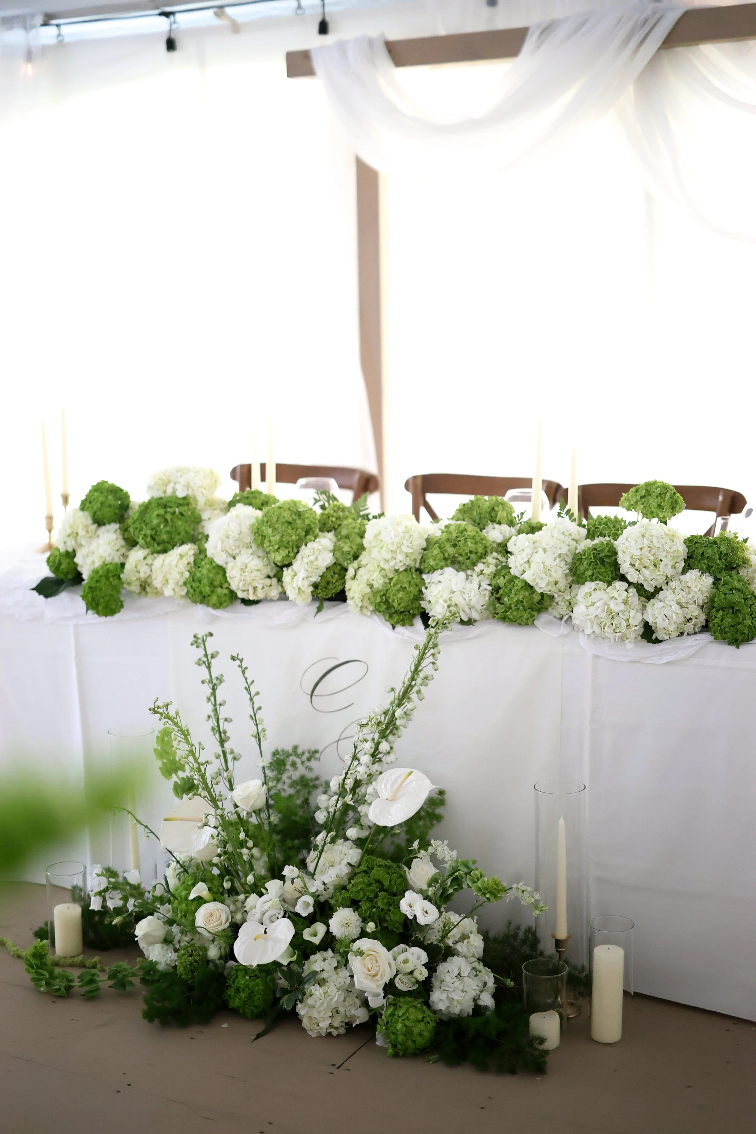 A wedding or event table decorated with white and green floral arrangements, candles, and draped fabric in a bright, airy setting.