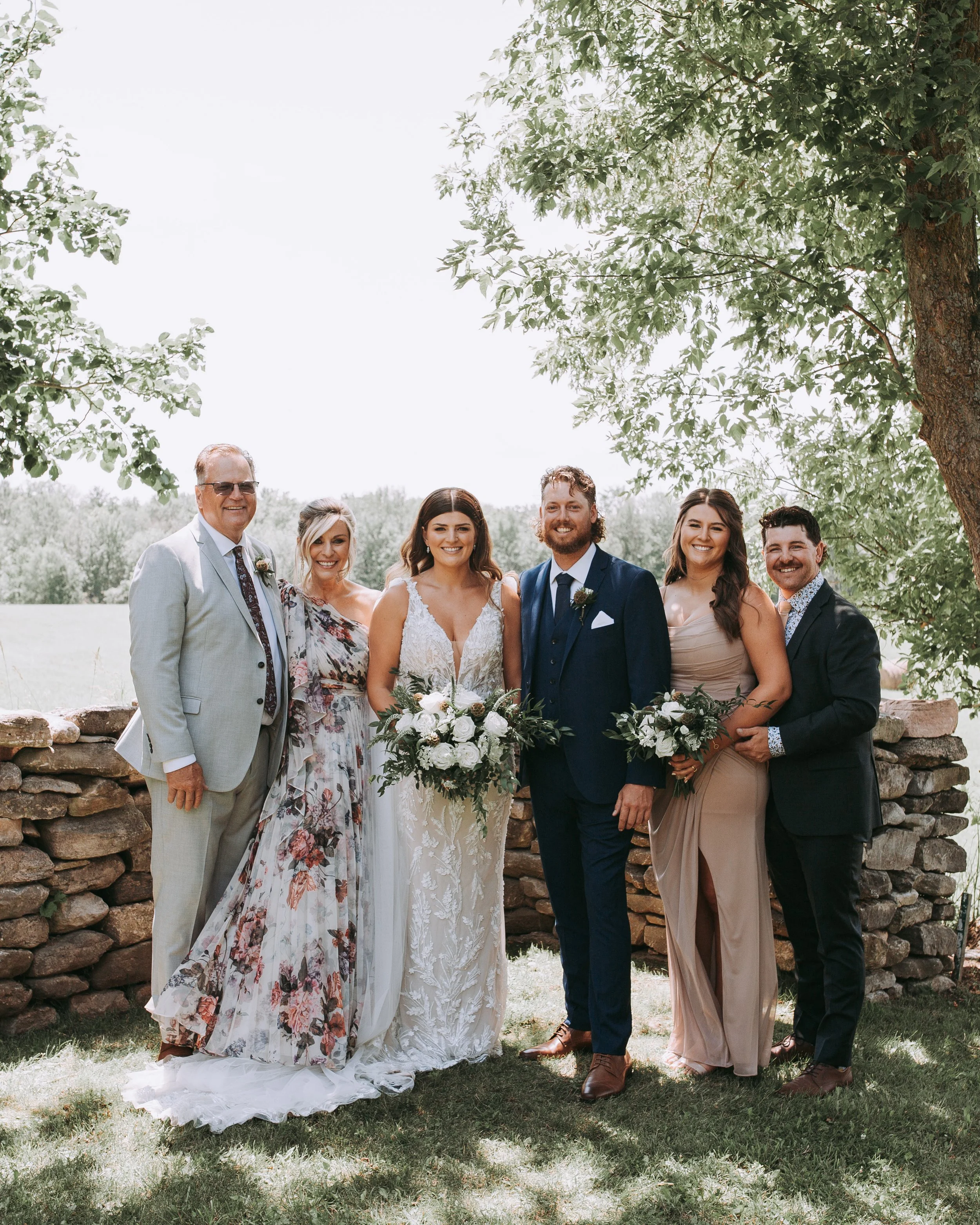 Group of seven people at a wedding outdoors, standing in front of a stone wall and a tree, smiling, with the bride holding a bouquet, and the groom standing next to her.