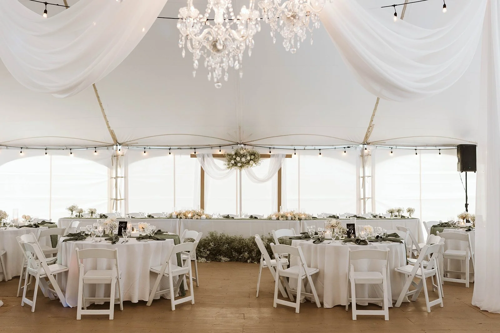 Elegant wedding reception tent with white round tables, white chairs, floral centerpieces, draped white fabric, a chandelier, string lights, and a central floral arrangement at the back.