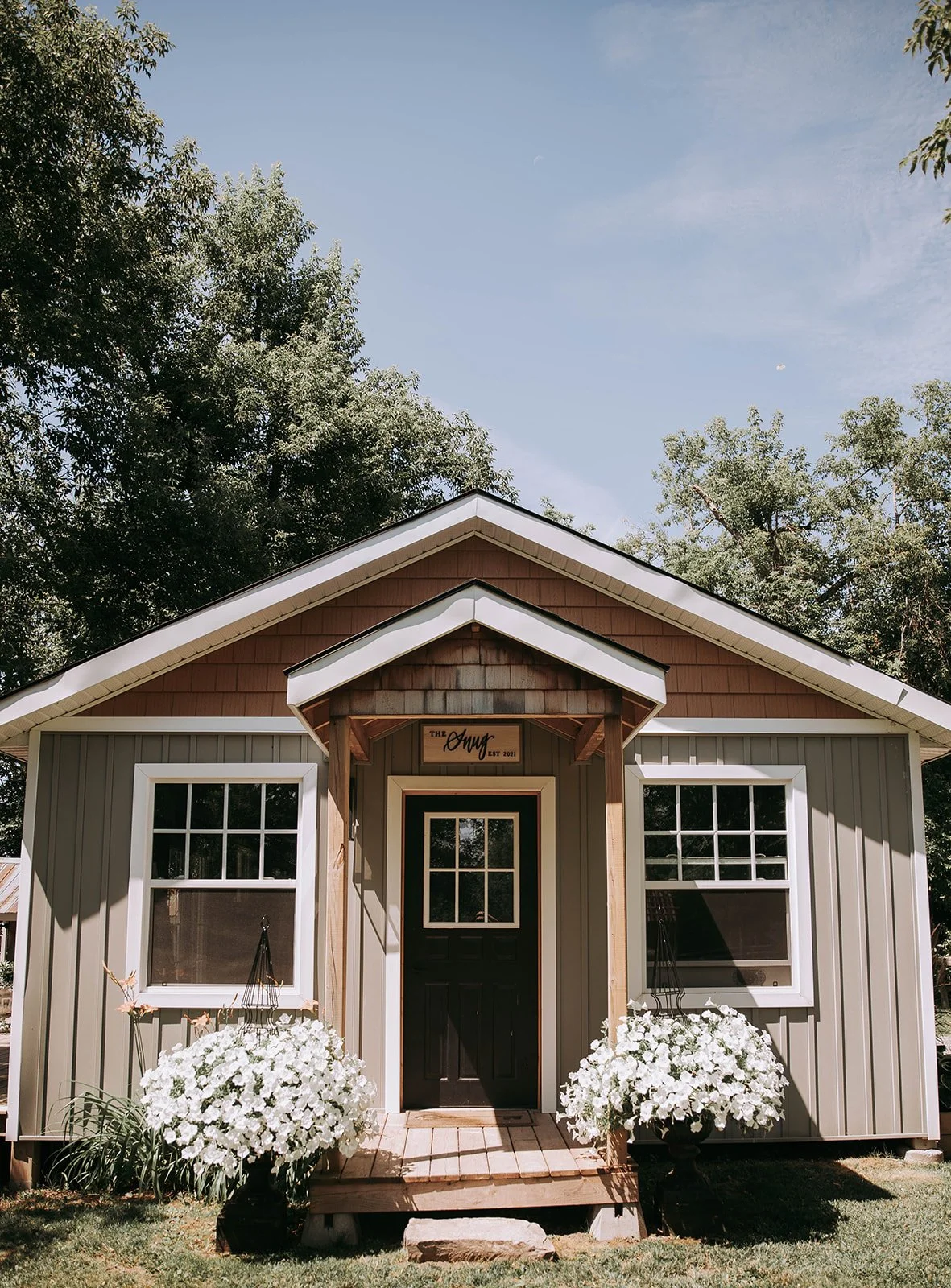 Front view of a small house or cottage with beige siding, white trim, and two large windows, surrounded by greenery and white flowers, with a wooden sign above the entrance reading "The Anny EST 2021".