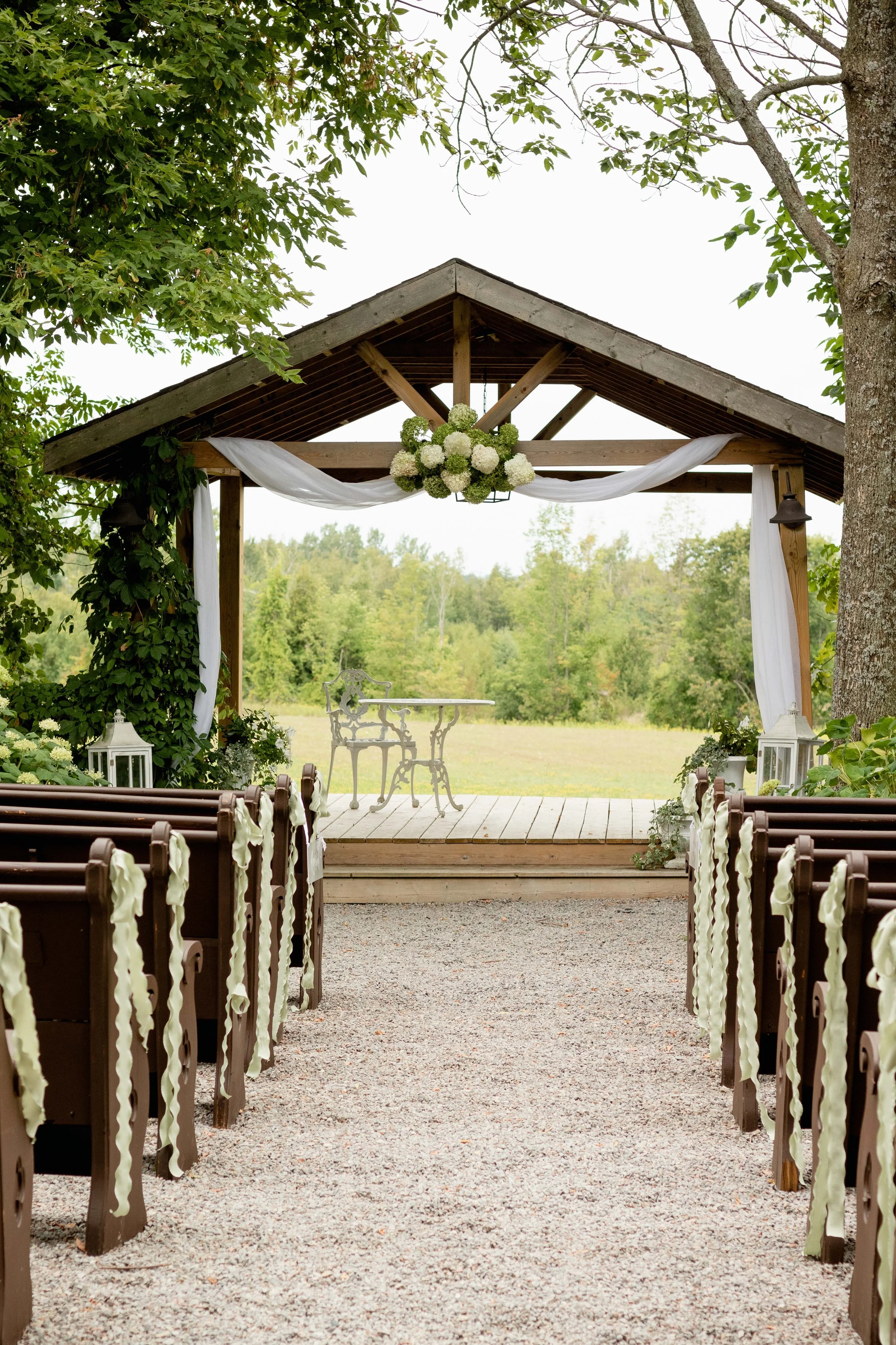 Outdoor wedding ceremony setup with a wooden altar decorated with white flowers and drapery, surrounded by benches with white ribbons, in a green outdoor setting.