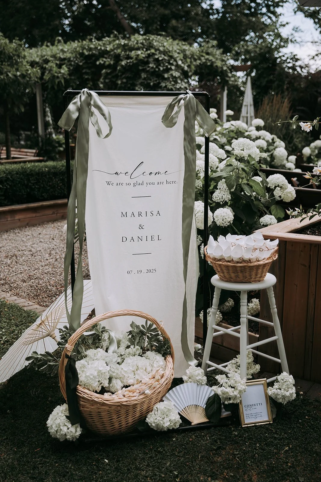 Wedding welcome sign with green ribbons, surrounded by white flowers, in an outdoor garden setting.