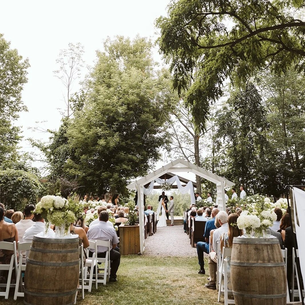 Outdoor wedding ceremony on a grassy area with guests seated in white chairs, floral arrangements, and a wooden arch under trees.