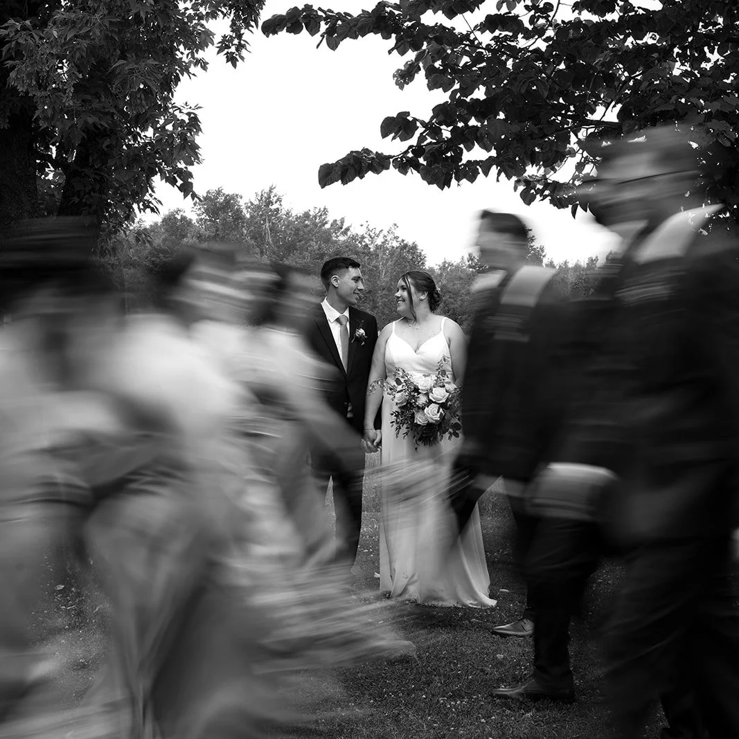 Black and white photo of a bride and groom holding hands, standing outdoors among trees, wedding guests blurred in motion around them.