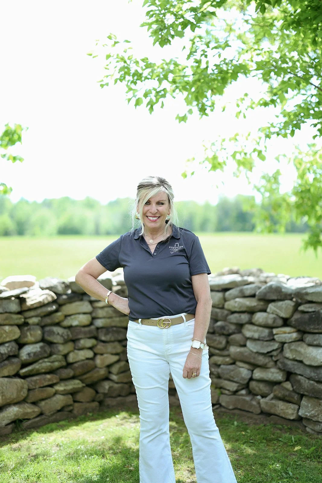 Woman standing outdoors beside a stone wall, smiling, with green trees and open field in the background.