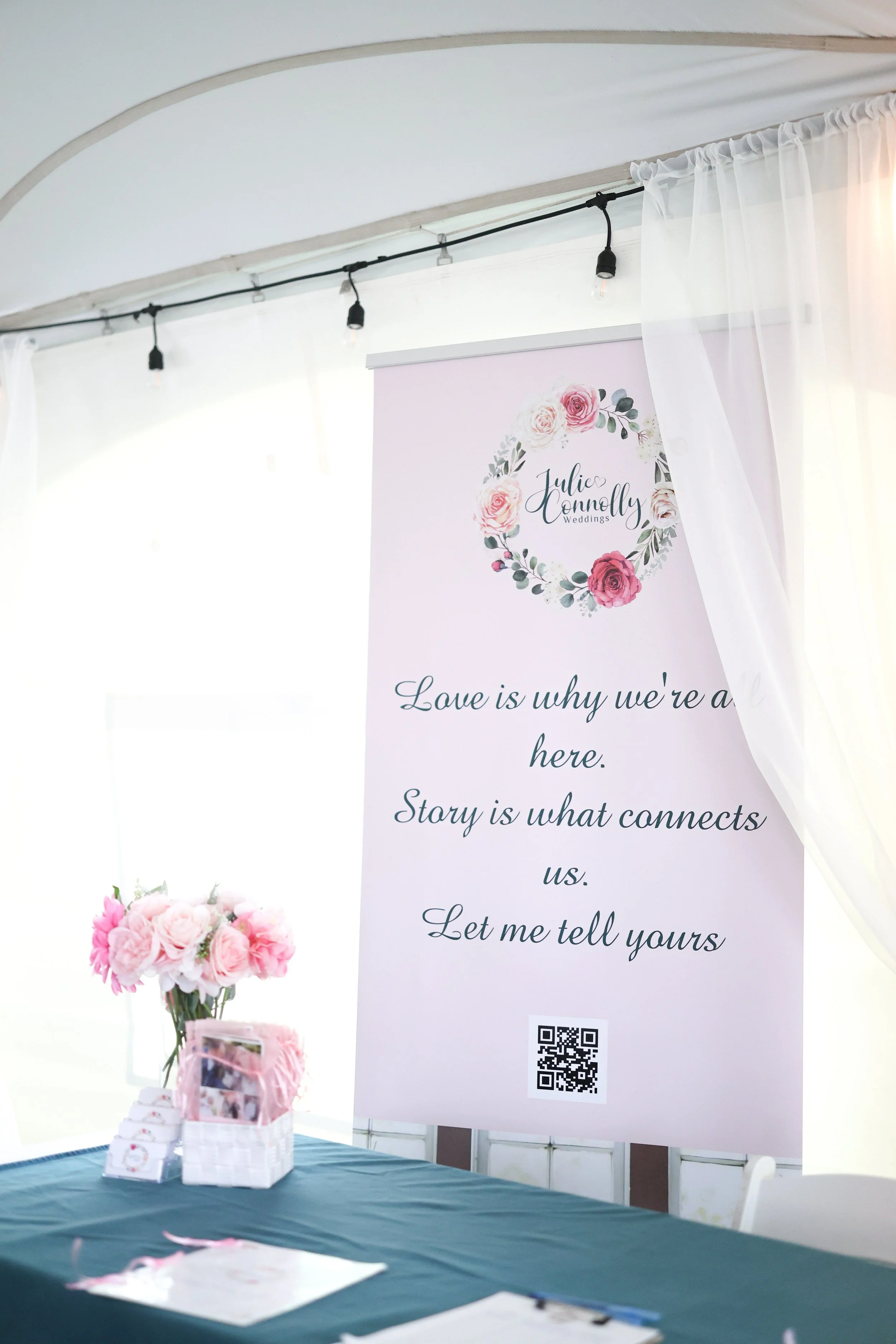 Wedding sign with pink floral wreath, reading, 'Julie Connolly Weddings. Love is why we're here. Story is what connects us. Let me tell yours.' A table with pink flowers, small boxes, and photos under a sheer curtain with string lights.