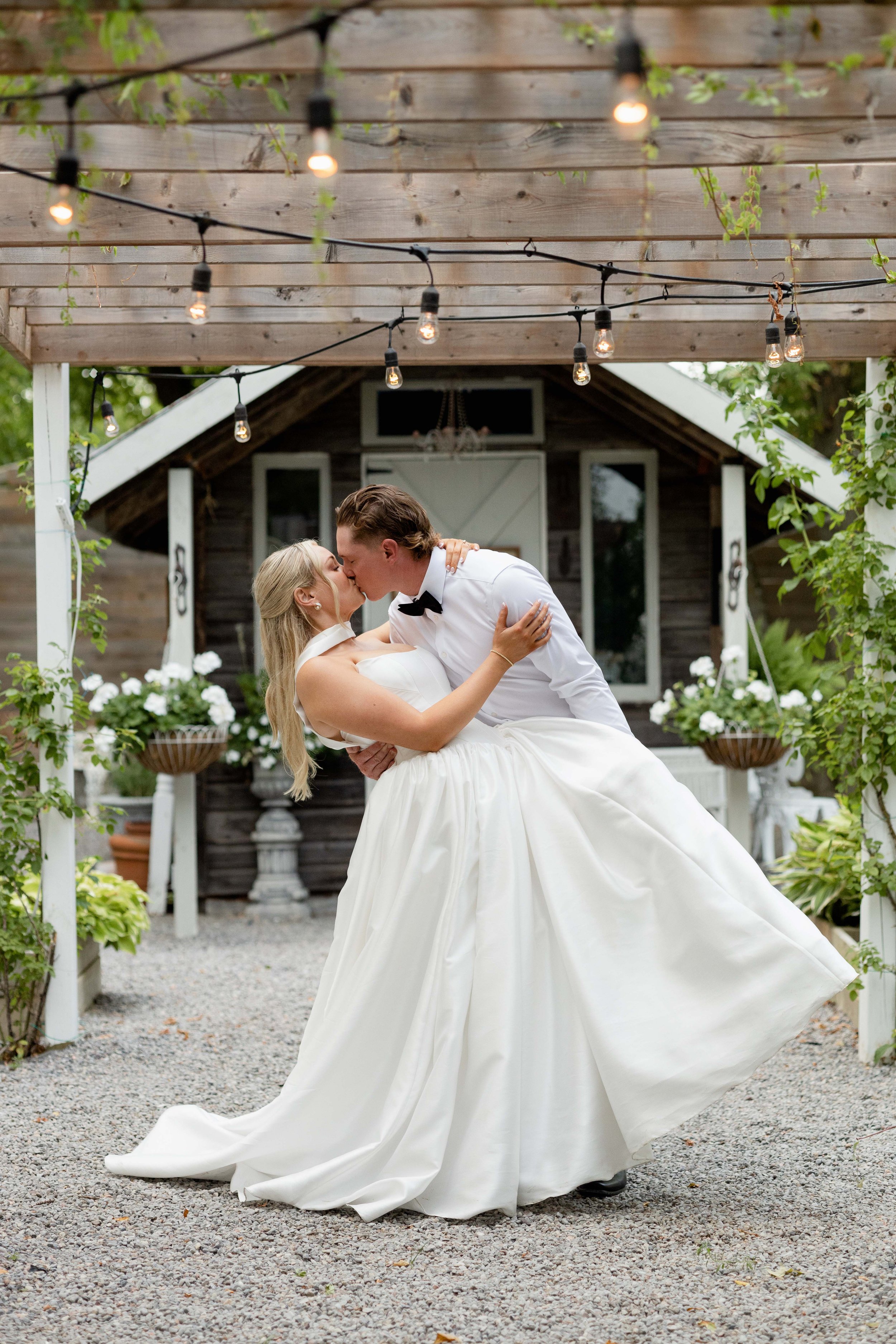 A bride and groom are sharing a kiss during their wedding dance outdoors under string lights, with a rustic barn and flower pots in the background.