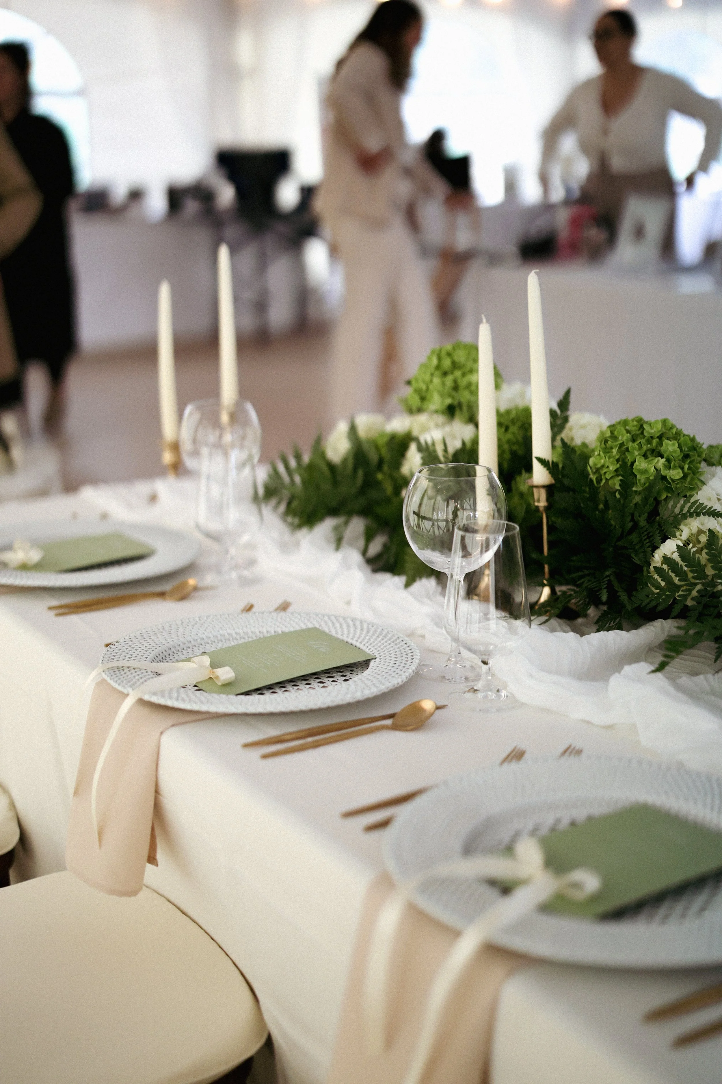 Elegant dining table setup with white plates, gold utensils, green menus, wine glasses, and a lush green floral centerpiece with white flowers and candles in the background.