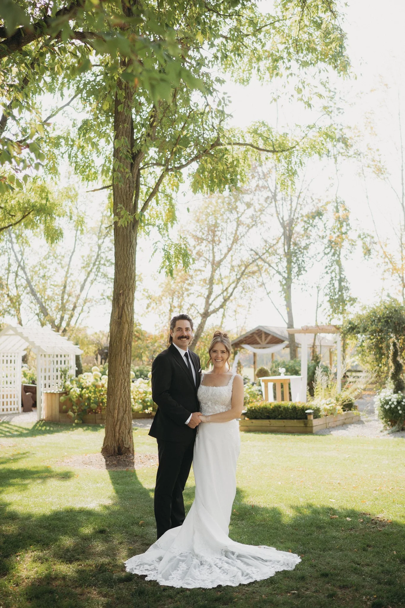 A newlywed couple, bride in a white wedding gown and groom in a black suit, posing together under a large tree in a garden setting on a sunny day.