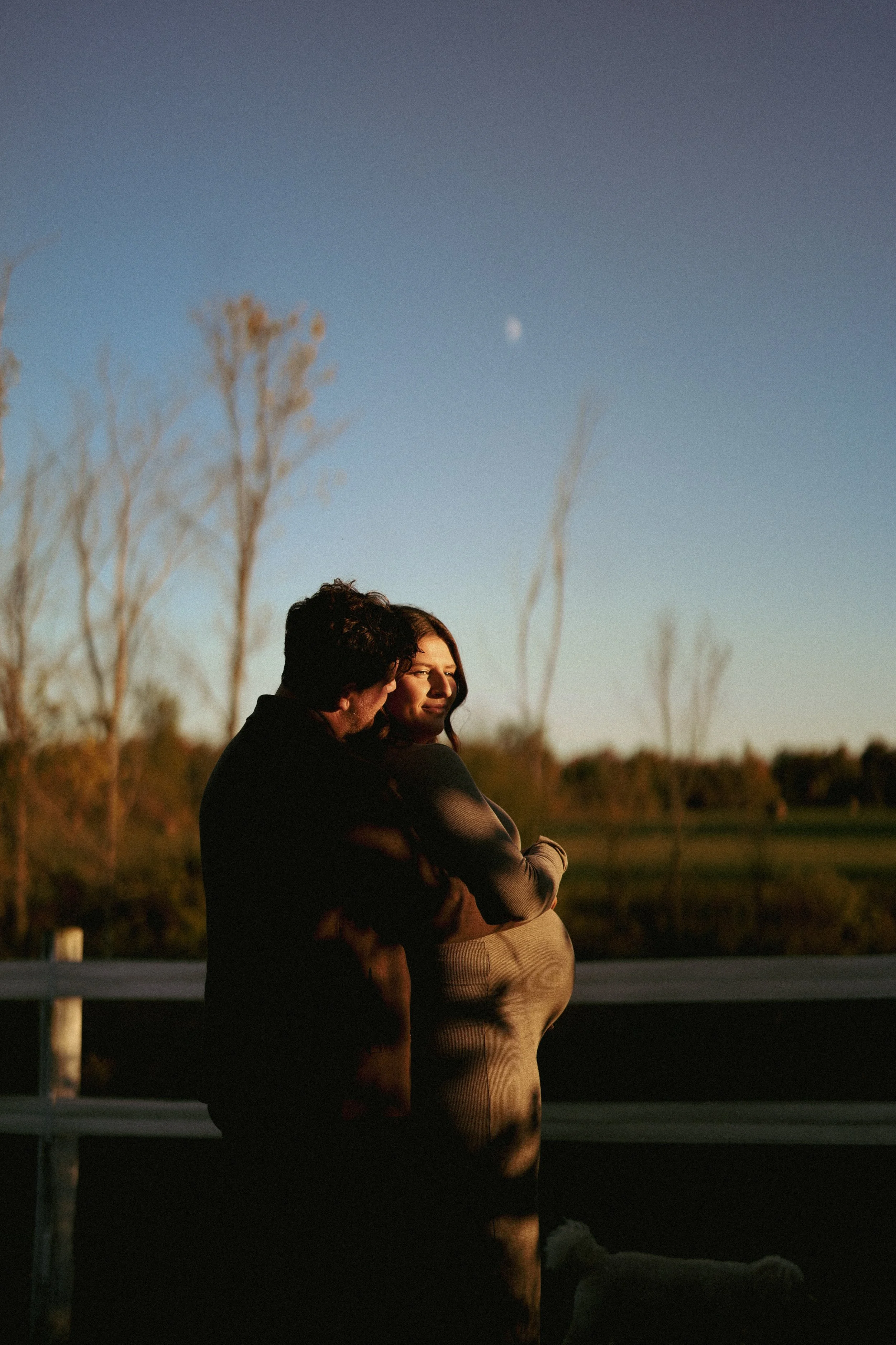 A couple standing on a bridge at sunset, with the woman noticeably pregnant and a dog nearby, against a blue sky with the moon visible.