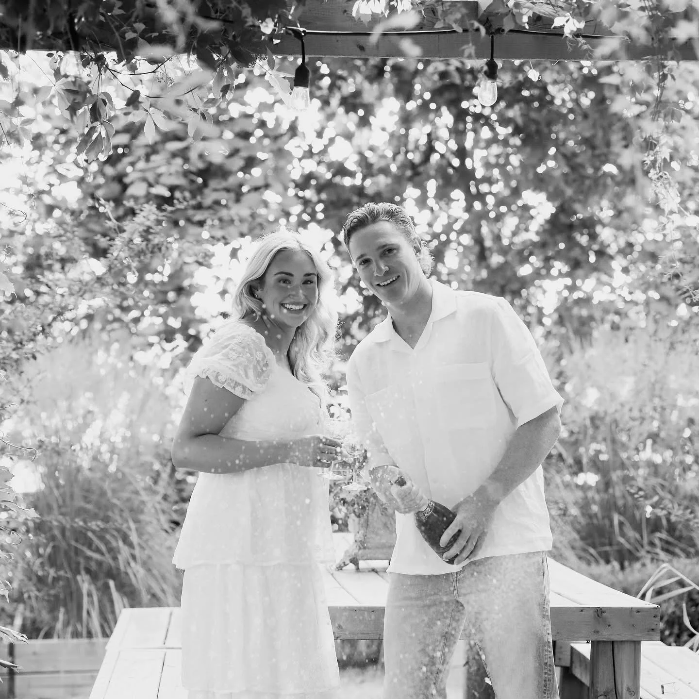 A black and white photo of a smiling woman in a white dress and man in a white shirt celebrating outdoors, pouring sparkling wine from a bottle.