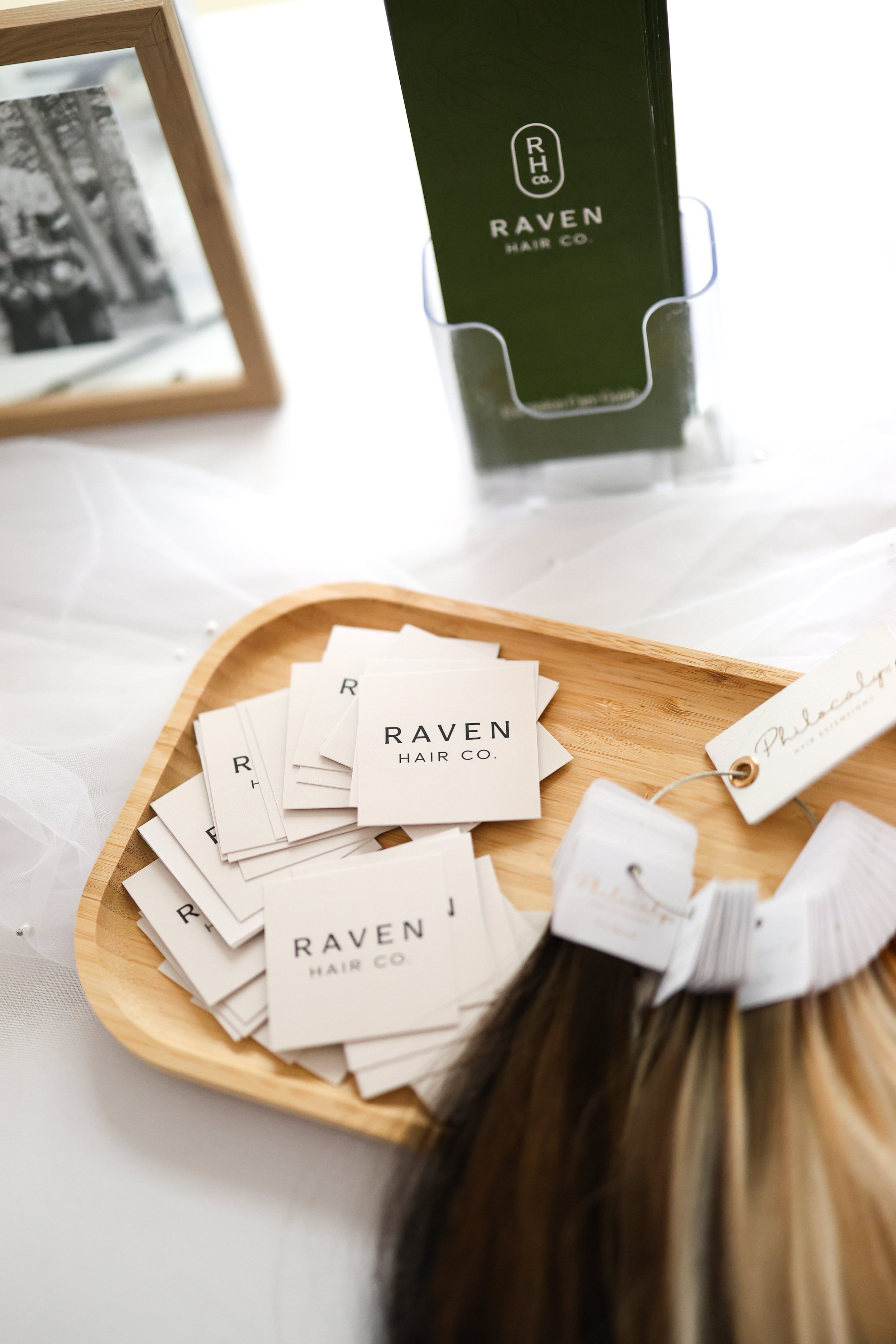 Display of business cards and a hair color swatch from Raven Hair Co. in a wooden tray on a white table, with a framed photo and a tall green box in the background.