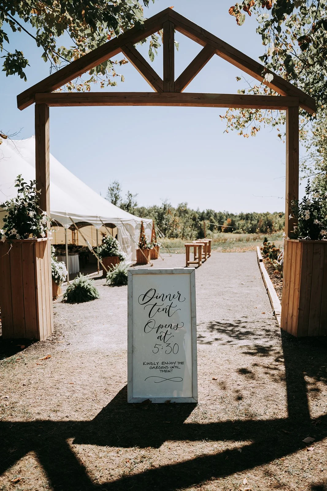 White signboard with black handwritten text indicating dinner tent opens at 5:30, placed at the entrance of an outdoor event space with a wooden archway, greenery, and floral decorations, under a clear blue sky.